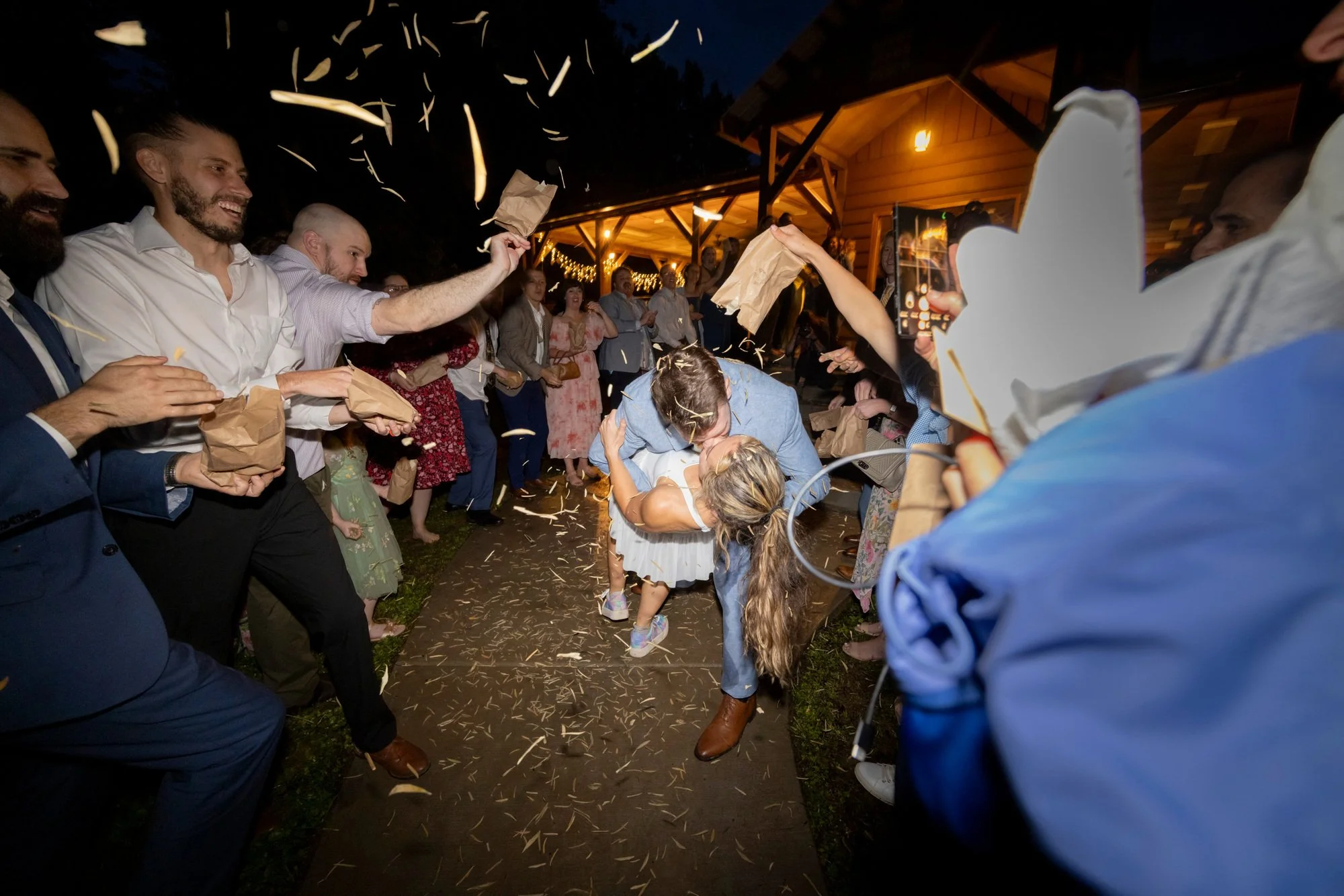 A group of people celebrate outside a wooden building at night, with confetti in the air. Some people are kissing on the ground while others watch and smile, capturing the moment on their phones.