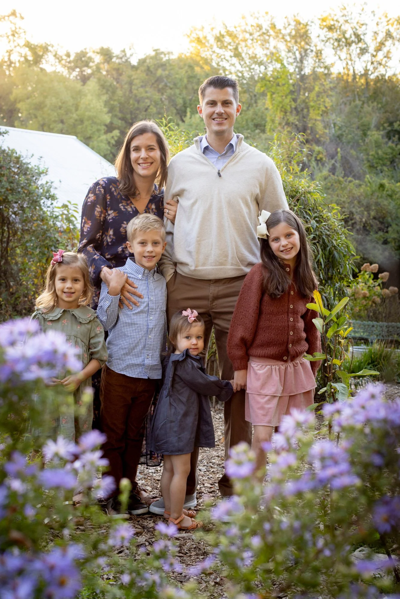 A family of seven, including two adults and five children, standing outdoors in a garden with trees in the background during daylight, smiling at the camera.