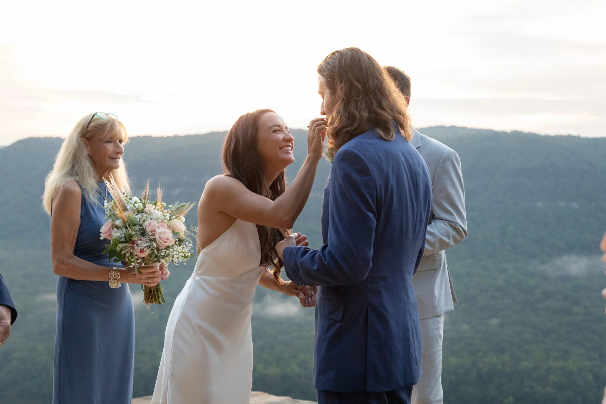 At an outdoor wedding ceremony, a bride with long brown hair, wearing a white dress, is smiling as she touches the groom's face. The groom, with shoulder-length brown hair, is dressed in a blue suit. An older woman with long blonde hair, wearing a bl