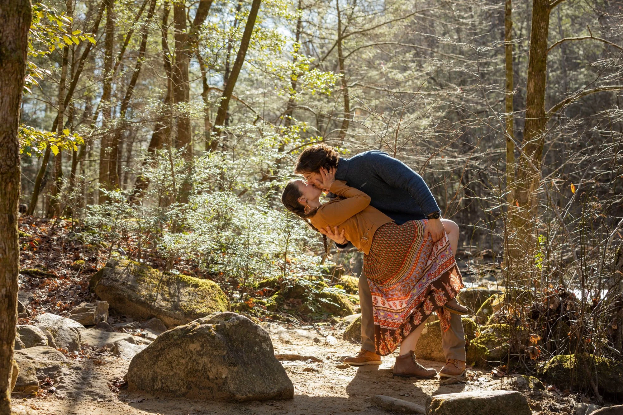 A woman in a patterned skirt and tan jacket being dipped by a man in a blue sweater in a forest, with sunlight filtering through the trees.