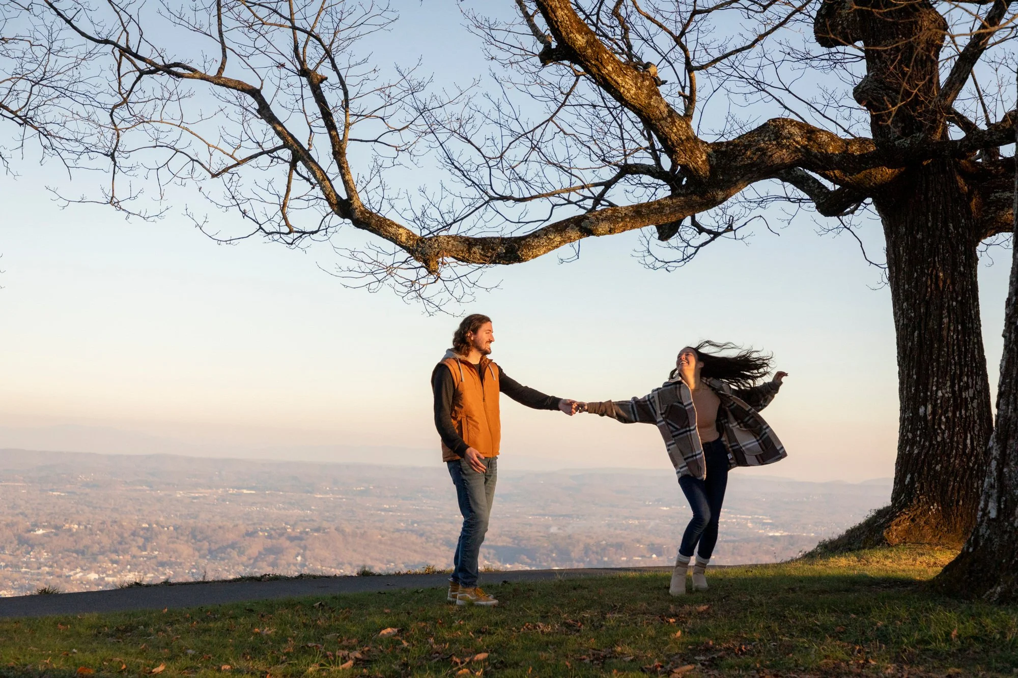 Two people, a man and a woman, are holding hands under a large leafless tree on a hilltop during sunset. They appear to be happy, with the woman spinning and the man smiling. The background shows a vast cityscape and mountains in the distance.