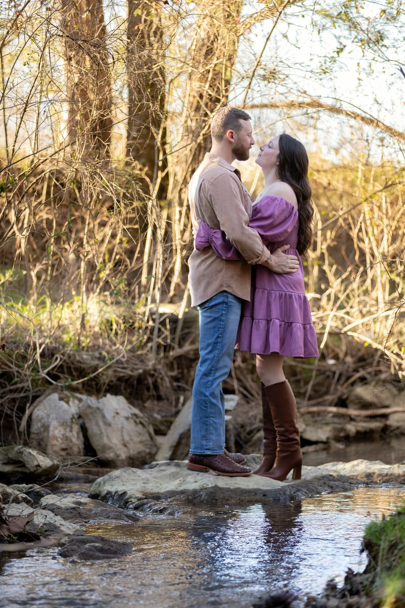 A couple embracing in a forest by a creek during sunset, both dressed in rustic clothing with the man wearing a tan shirt and jeans, and the woman in a purple dress and brown boots, standing on rocks in the shallow water.