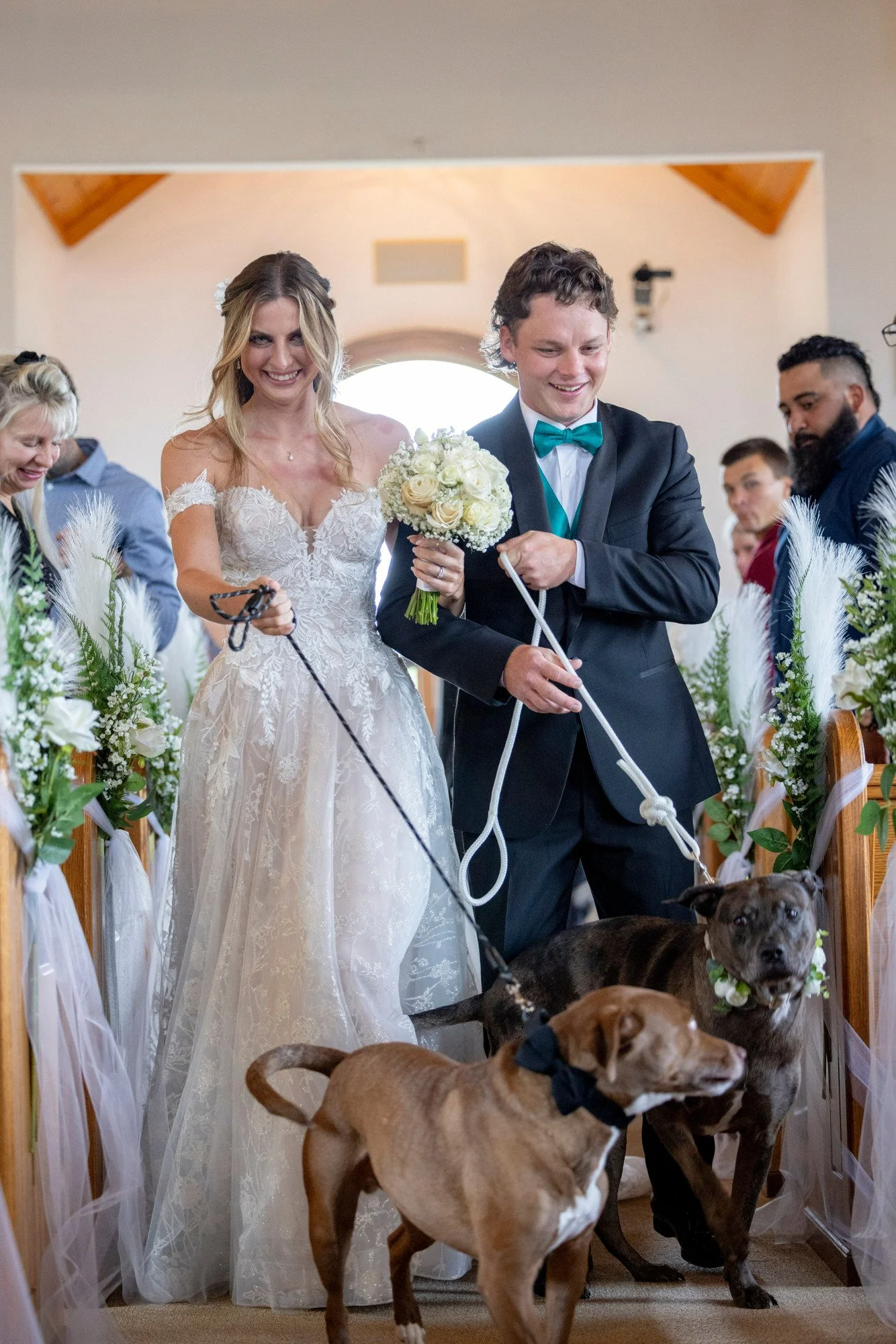 A bride and groom walking down the aisle with multiple dogs during a wedding ceremony inside a church or chapel. The bride is wearing a white lace gown, and the groom is dressed in a black suit with a turquoise bow tie. The aisle is decorated with wh