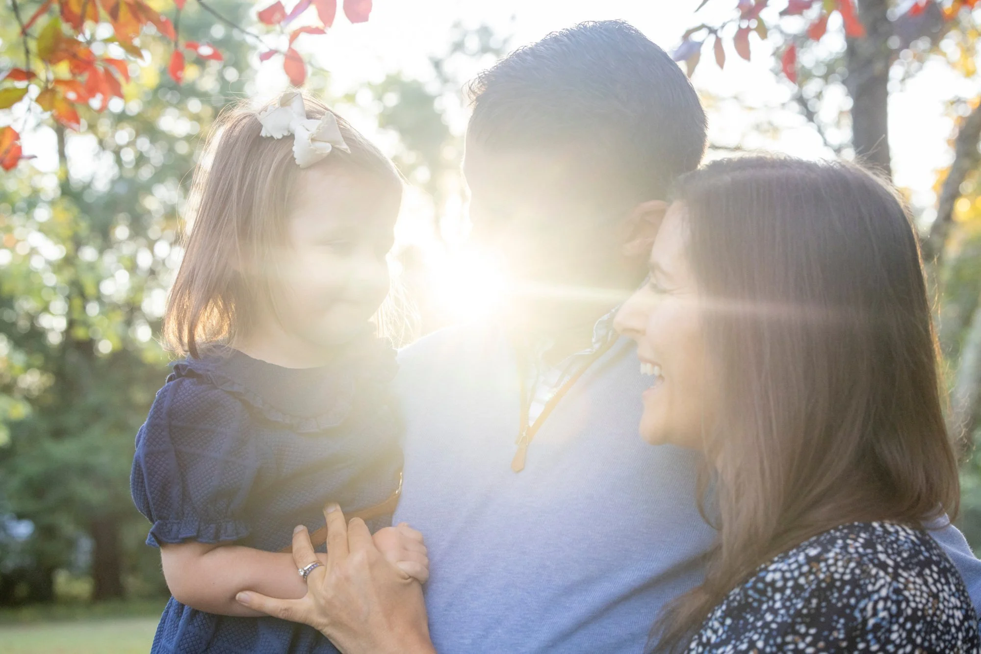 Family portrait outdoors with sunlight shining through trees, showing two women and a young girl smiling and interacting.
