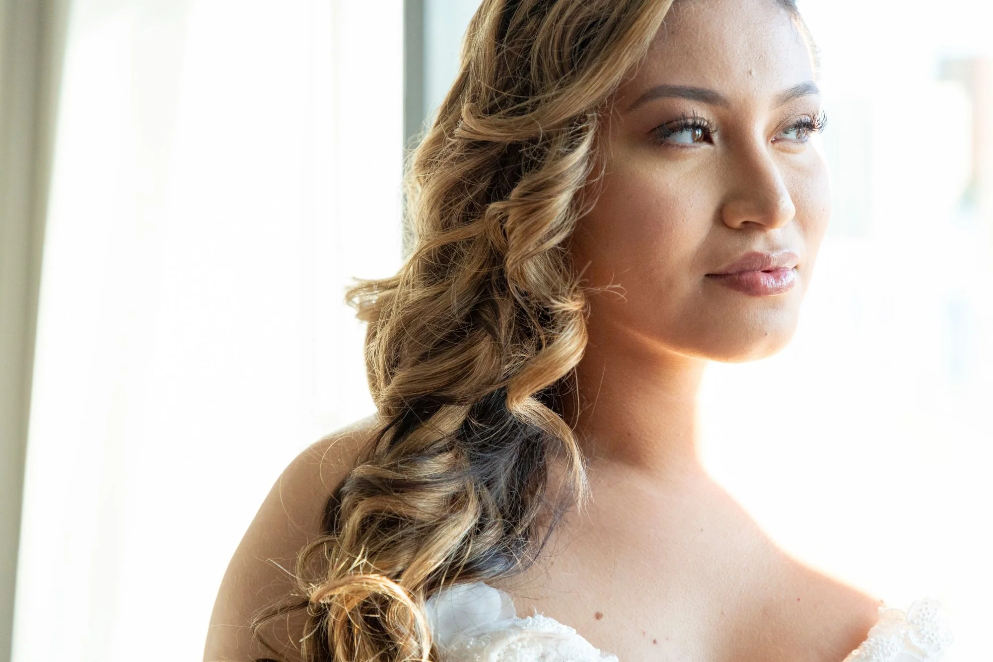 Close-up of a woman with styled curly hair, looking out a window, with natural light illuminating her face.