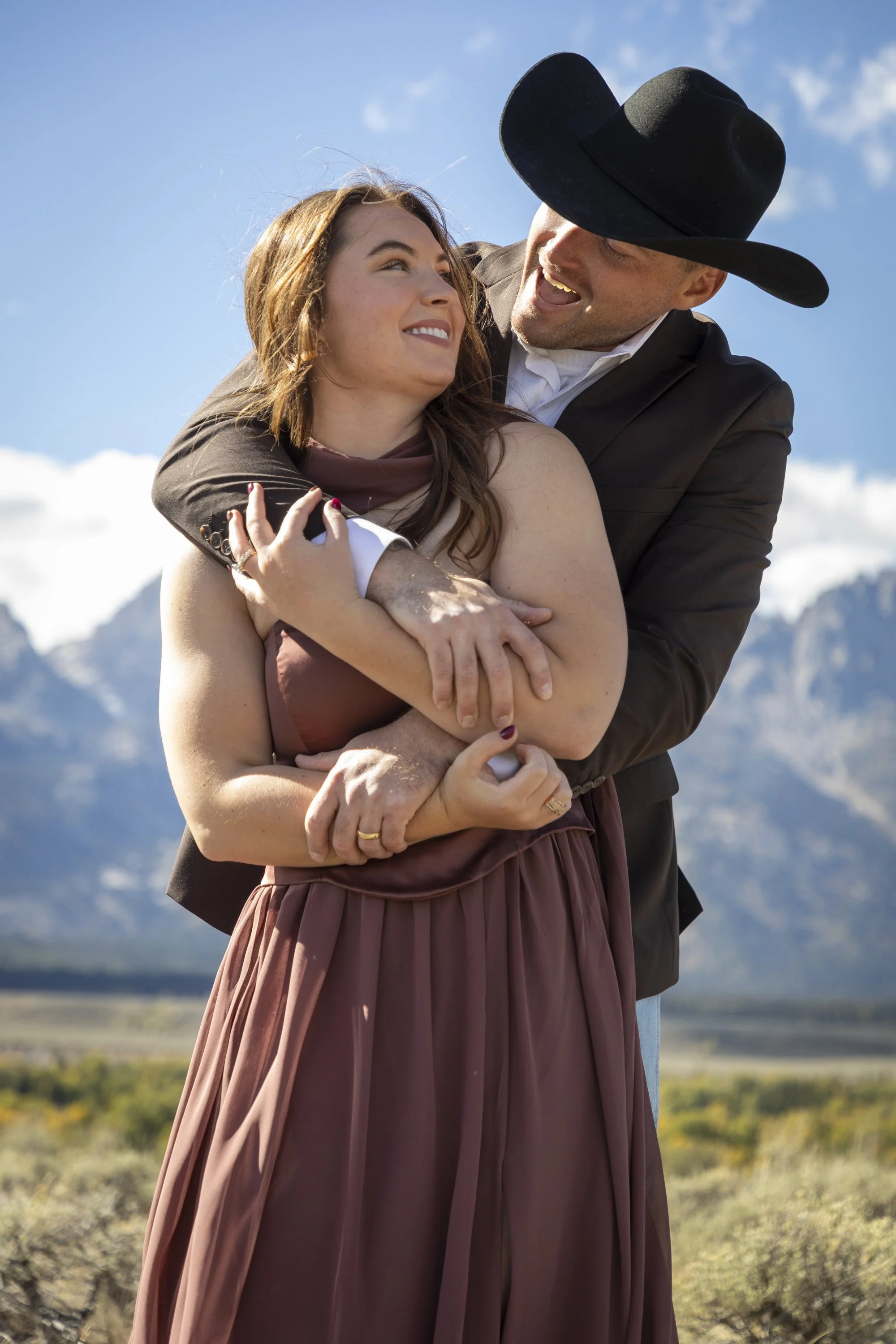 A couple embracing outdoors with mountains and a blue sky in the background. The man is wearing a black hat and a suit, smiling, while the woman has shoulder-length hair and is smiling back at him.