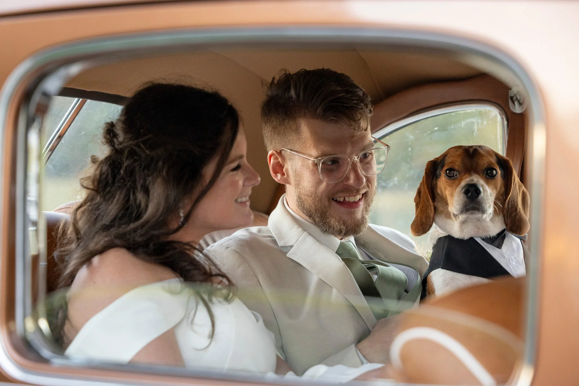 A happy couple and a dog in a tuxedo sitting inside a vintage car, seen through a car window.