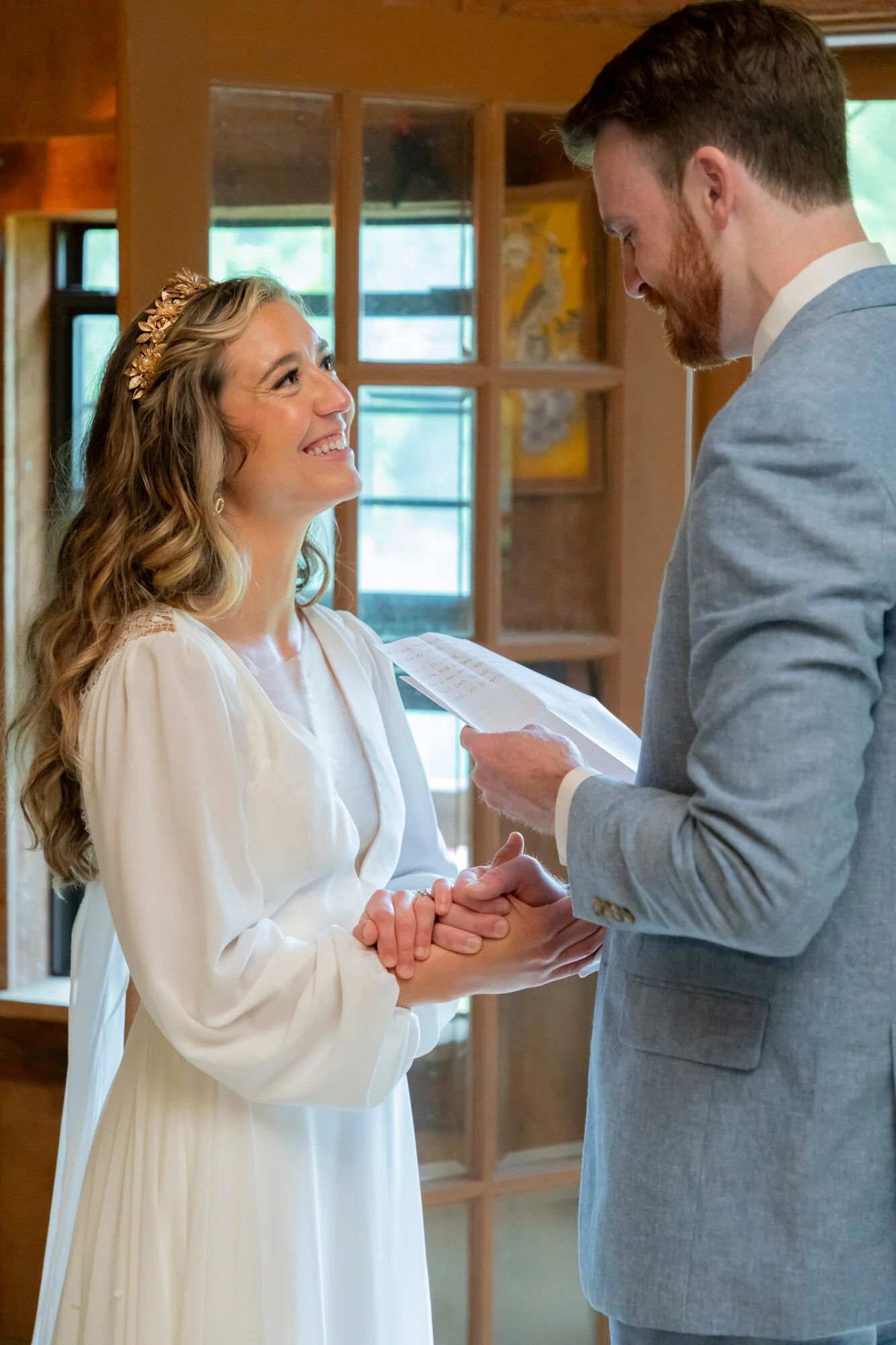 A bride and groom holding hands during their wedding ceremony, with the bride smiling at the groom as he looks down at her, holding a piece of paper or vows.