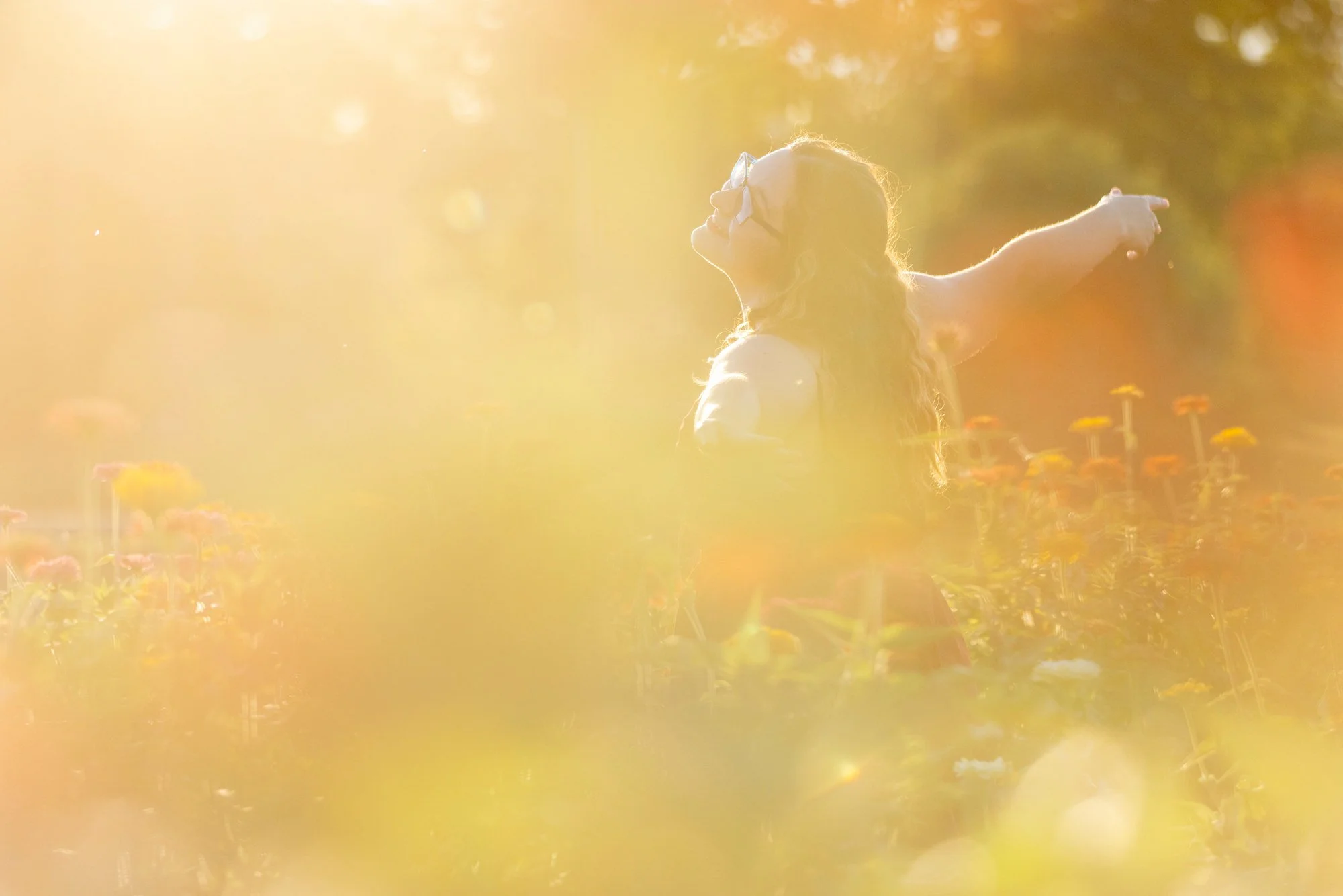 A young woman with long hair and glasses standing in a field of flowers, smiling and pointing into the distance as sunlight creates a warm, glowing atmosphere.