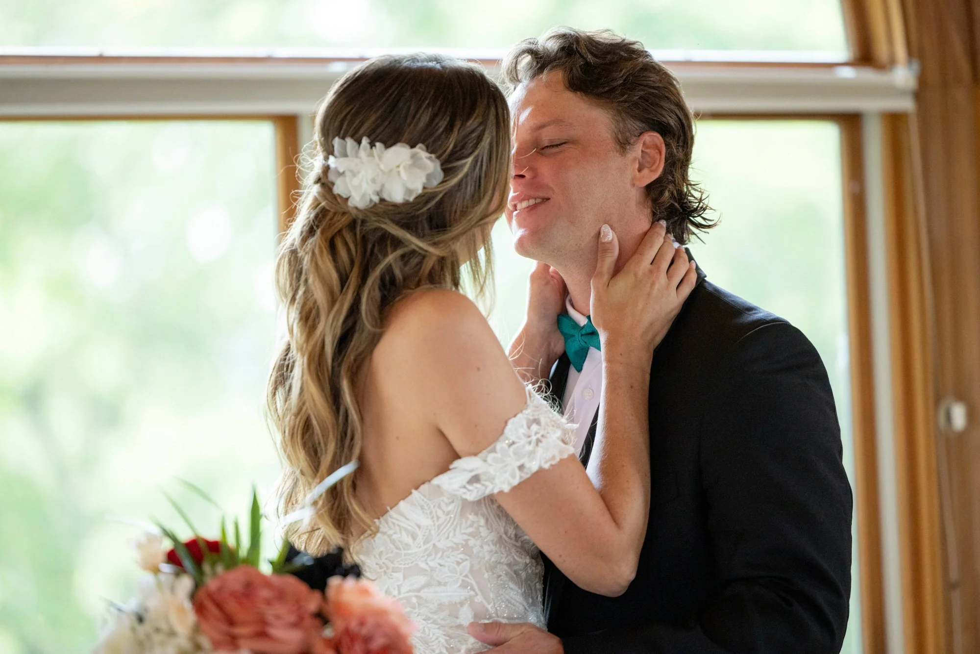 A bride and groom share a close moment, smiling and touching foreheads, during their wedding ceremony in a room with large windows and natural light.