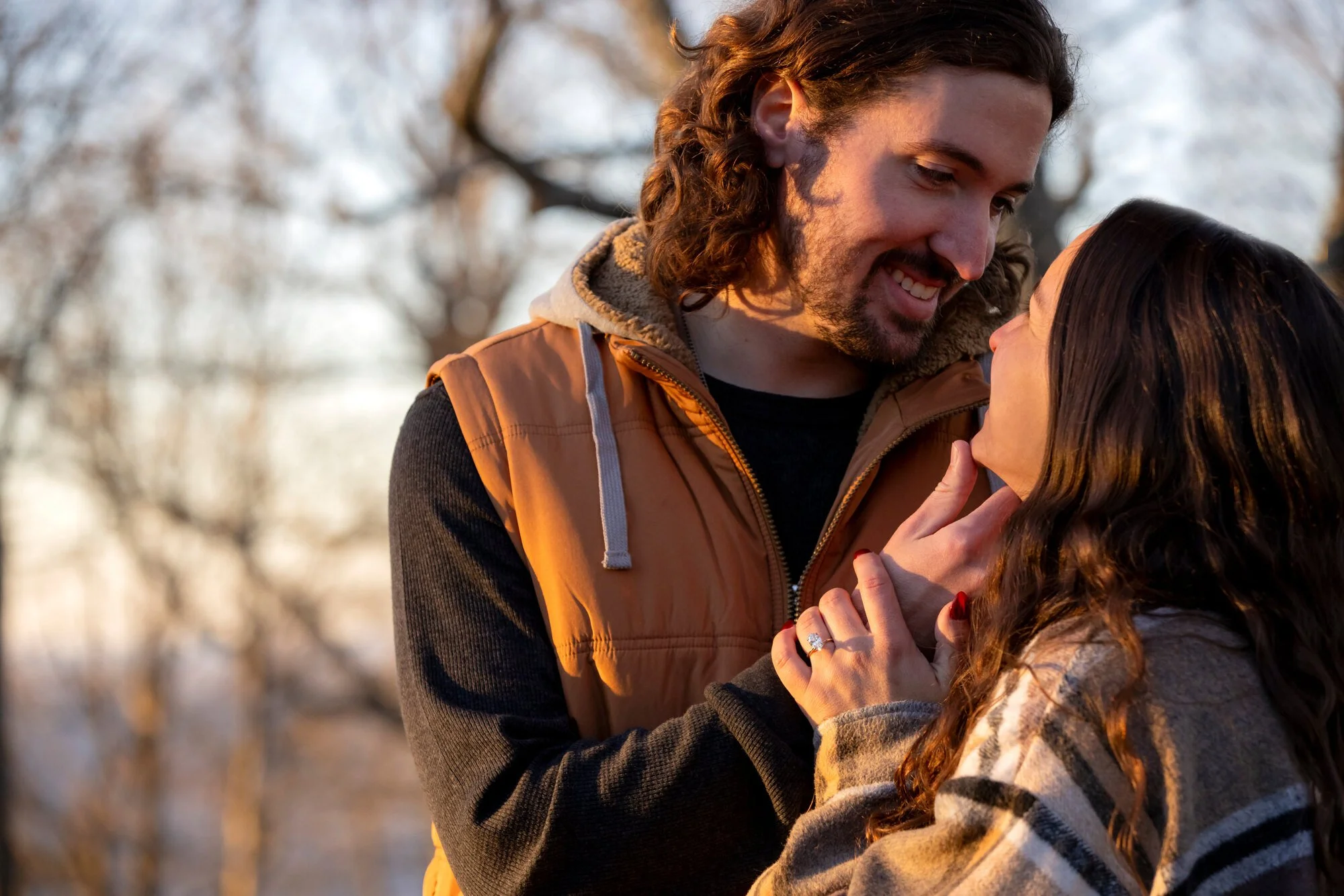 A man with long brown hair, a beard, and wearing a brown vest is smiling and looking into the eyes of a woman with long brown hair and red nails, who is looking up at him. They are outdoors with blurred trees in the background, during sunset or sunri