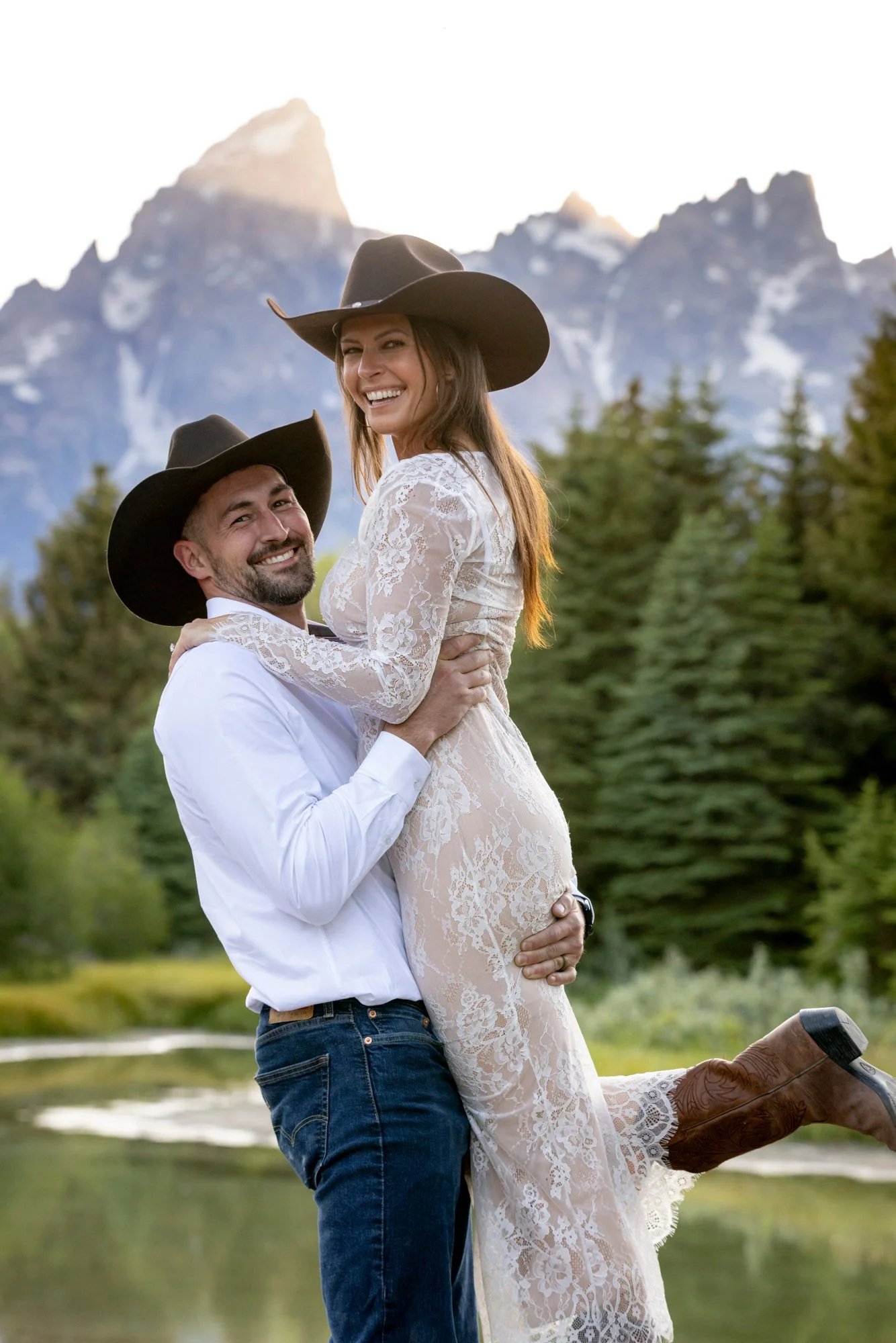 A man wearing jeans and a white shirt lifts a woman in a lace dress and cowboy boots with mountains in the background.