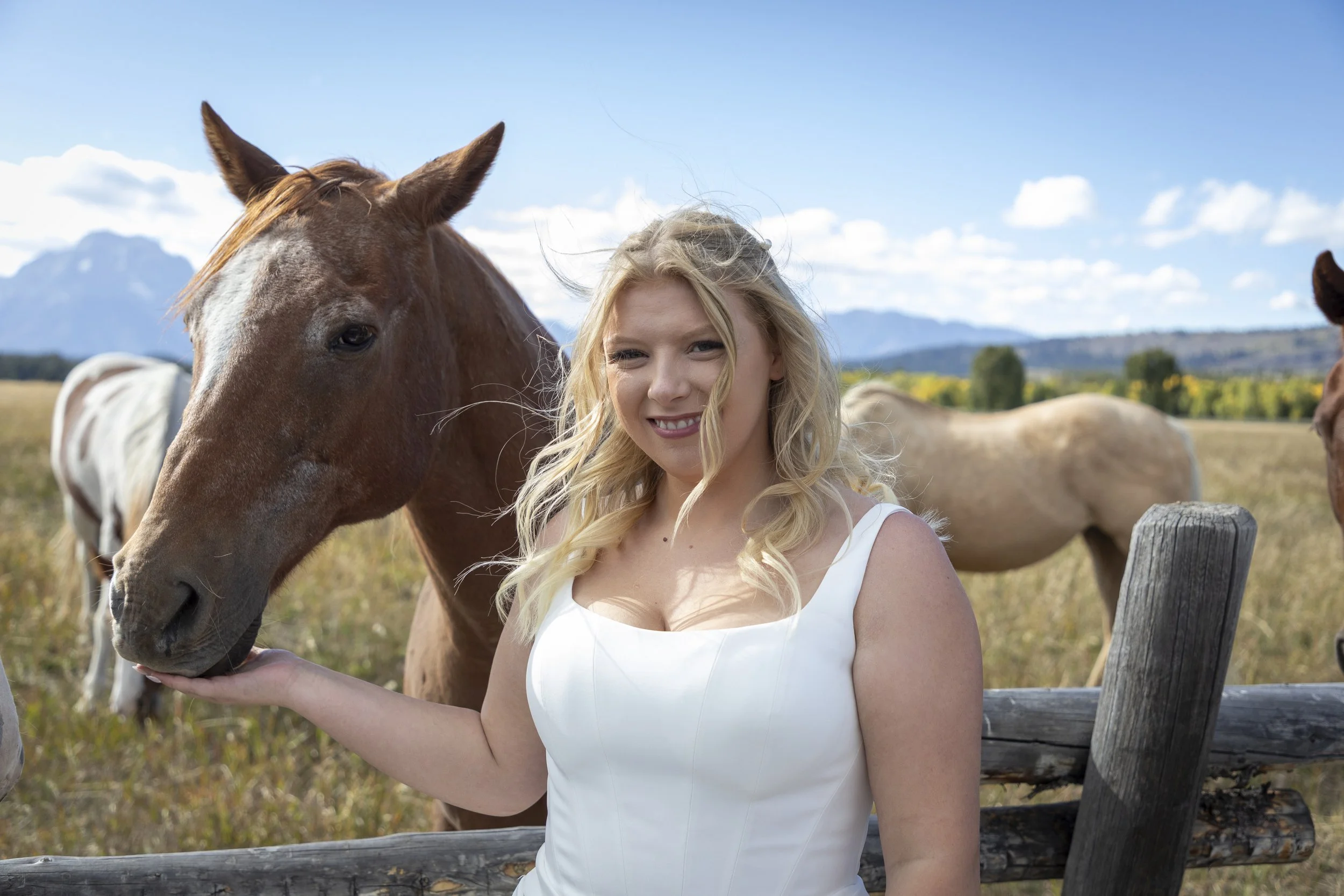 A young woman with curly blonde hair and a white dress standing near a wooden fence, petting a brown and white horse in a rural landscape with mountains and trees in the background.