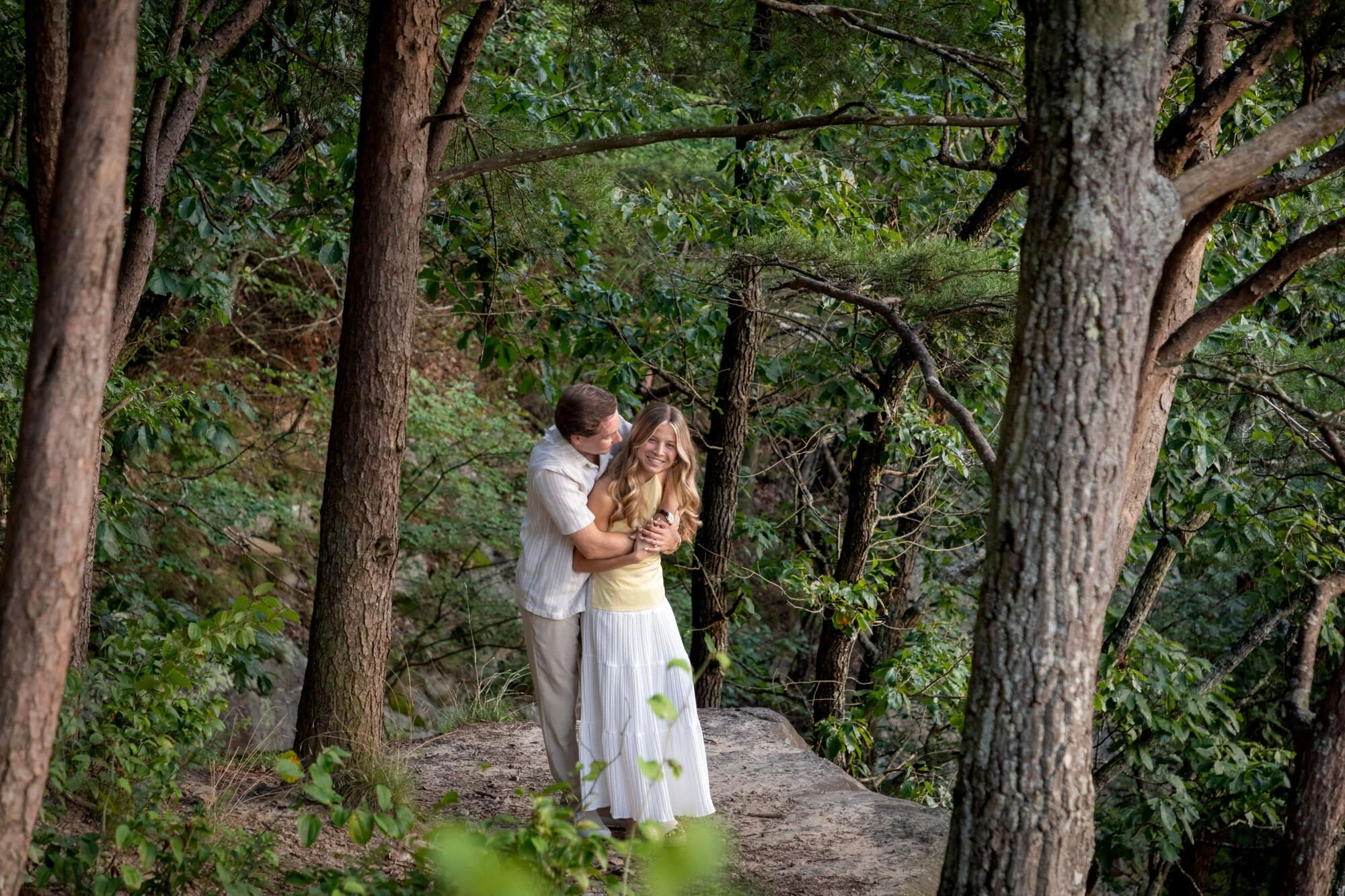 A couple standing on a forest trail, embracing each other, surrounded by tall trees and green foliage, with the woman smiling.