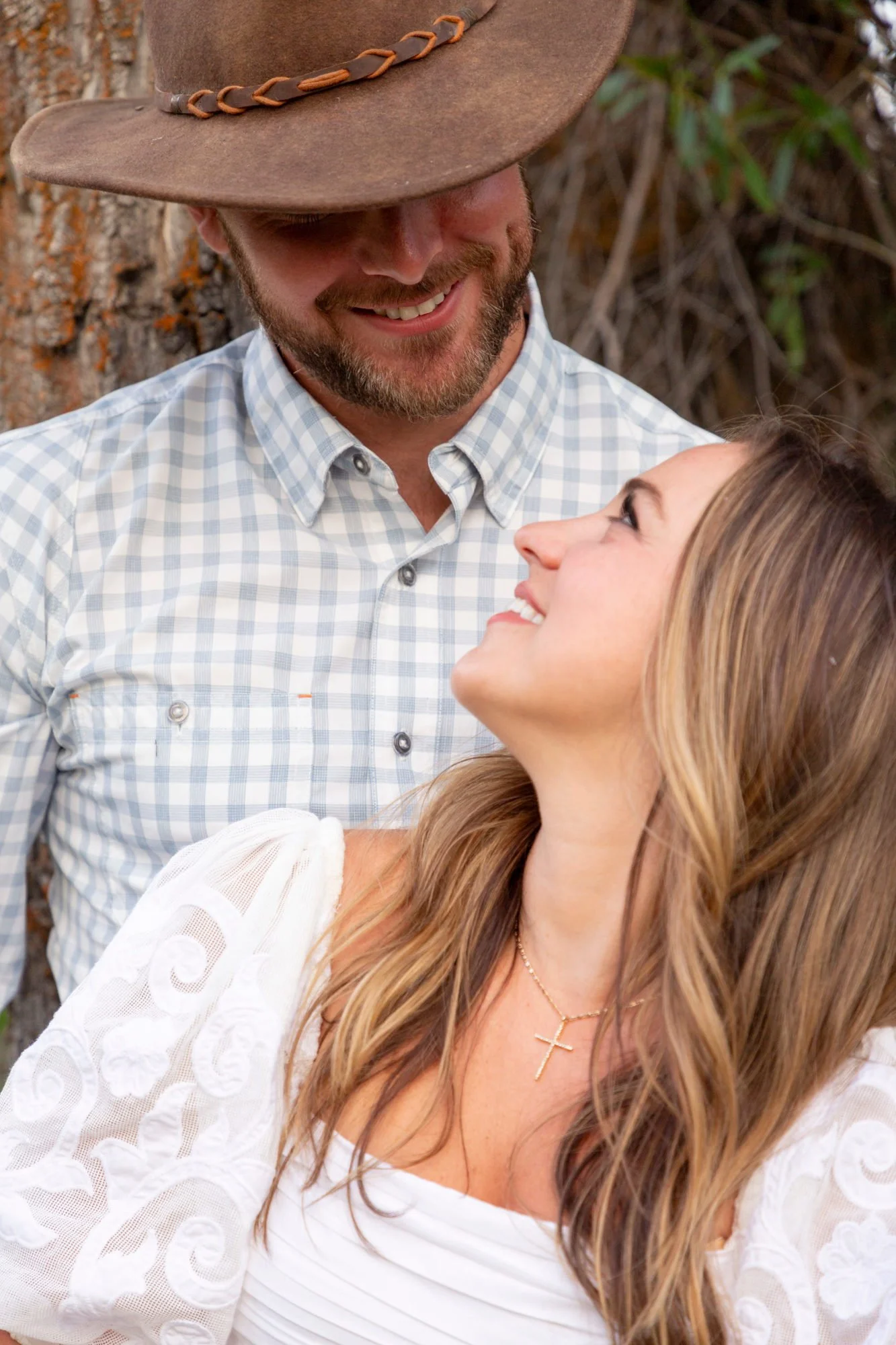 A man wearing a cowboy hat and a woman with long, wavy hair smiling at each other outdoors near a tree.