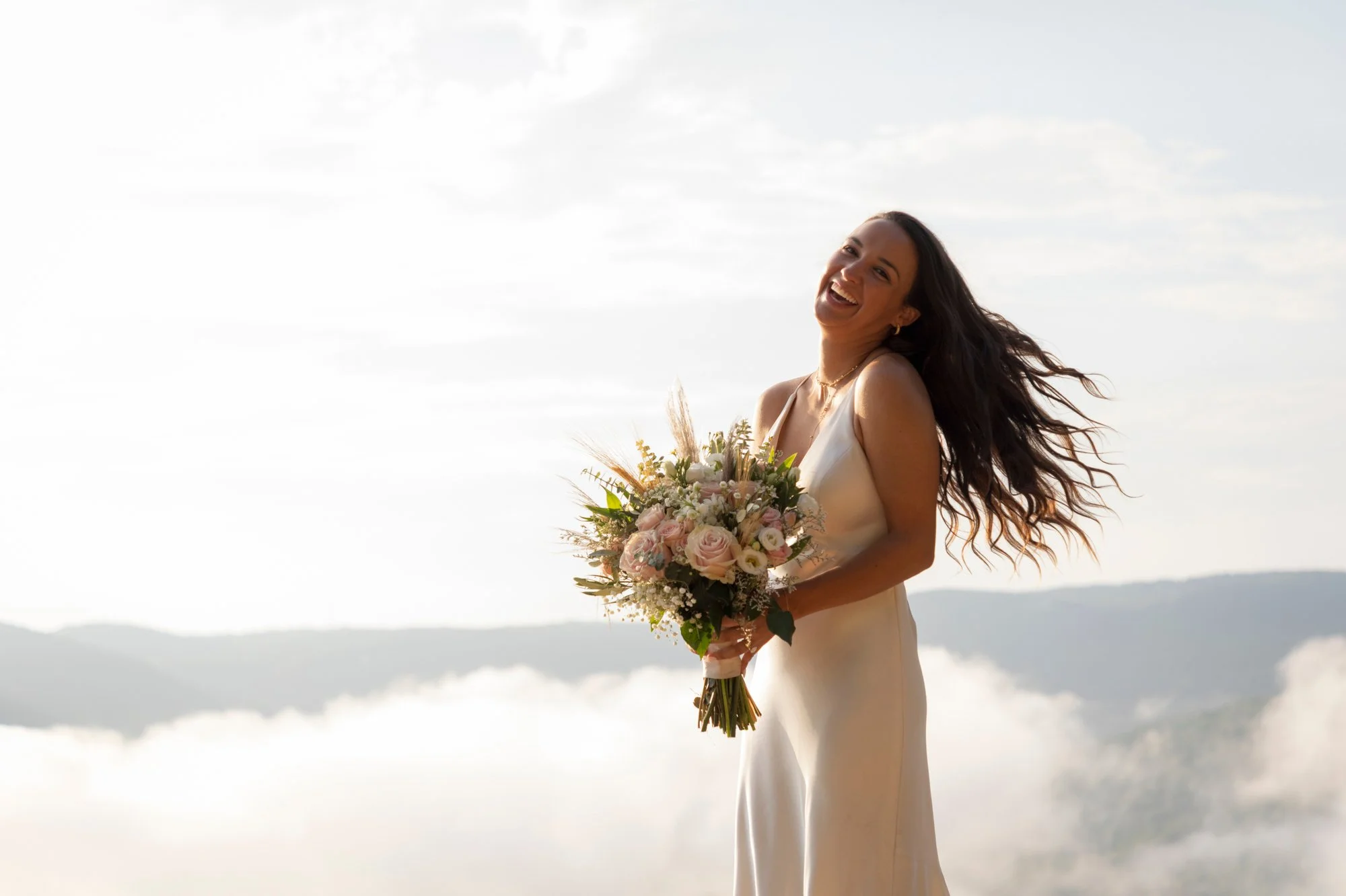 A woman in a white dress holding a bouquet of pink and white flowers, smiling outdoors with a bright sky and distant mountains.