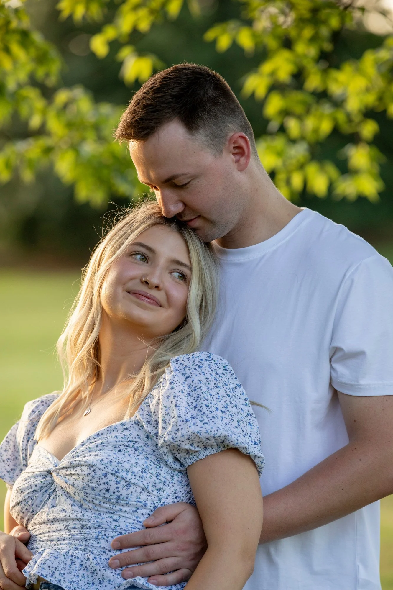 A young couple embraces outdoors, with the man gently resting his forehead on the woman's head, both with serene expressions, amid a green, leafy background.