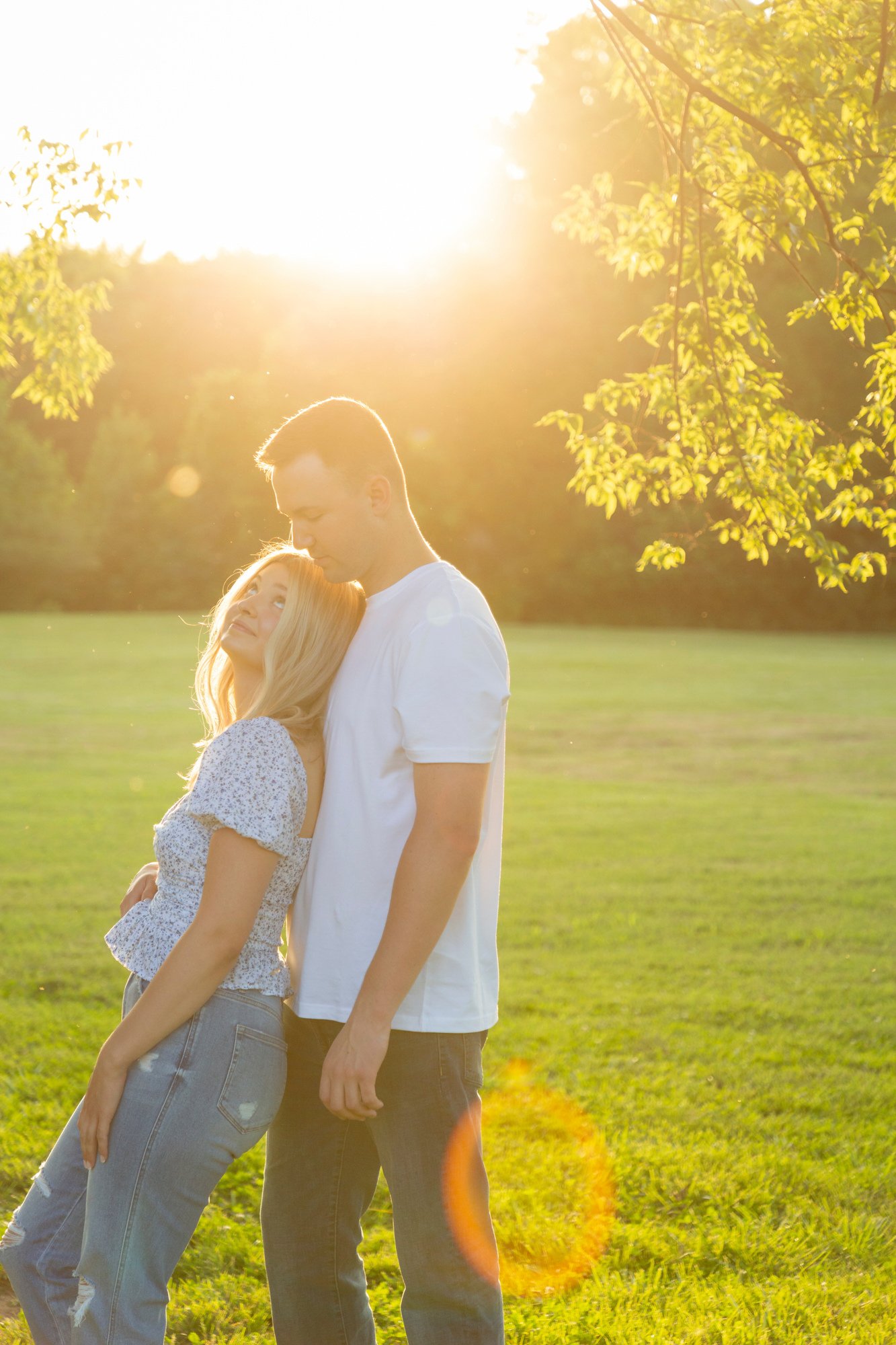 A young man and woman standing close together outdoors in a sunlit park, with the sun shining behind them and trees with green leaves in the background.