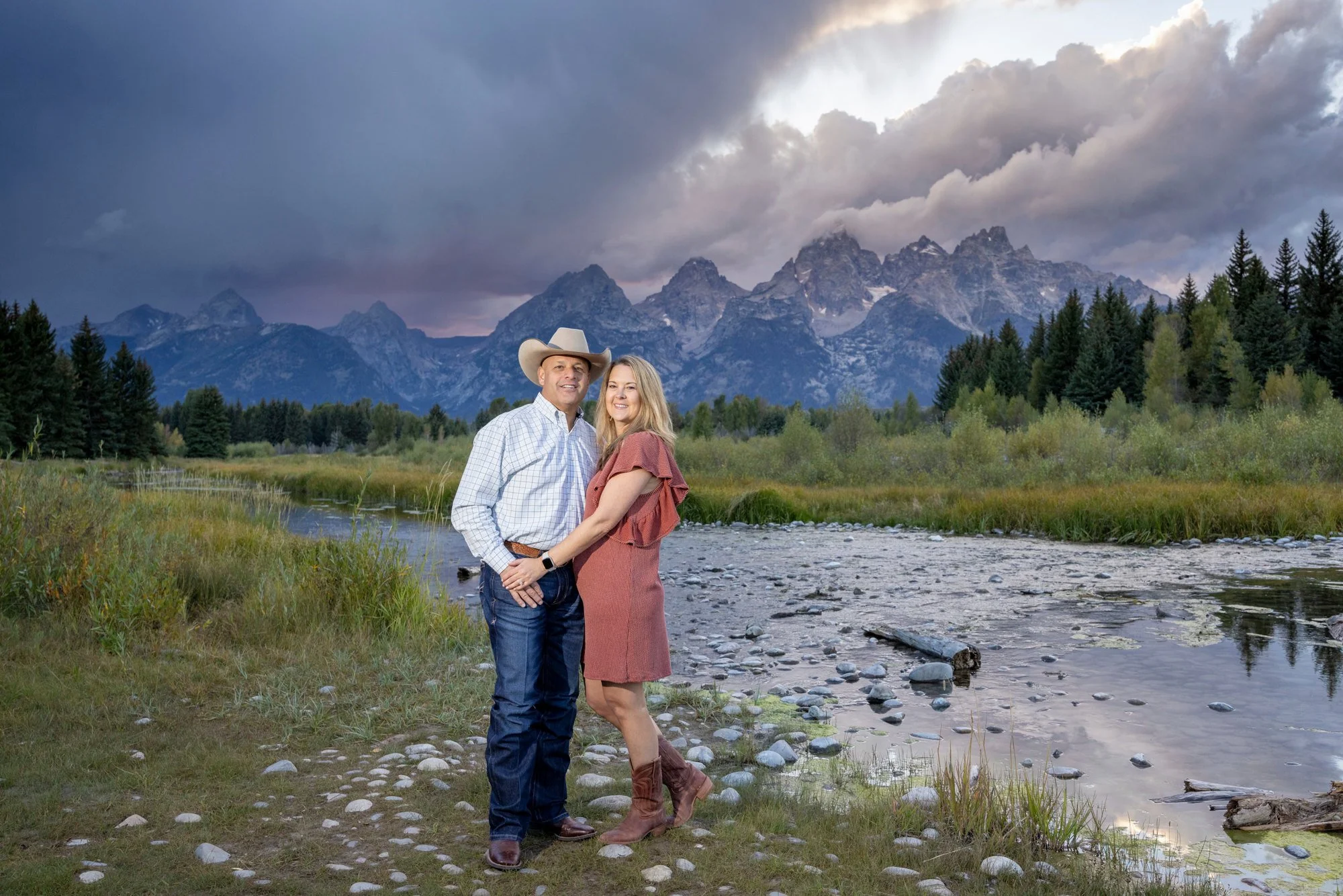 A couple standing close together on a rocky riverbank with mountains and a cloudy sky in the background, smiling at the camera.