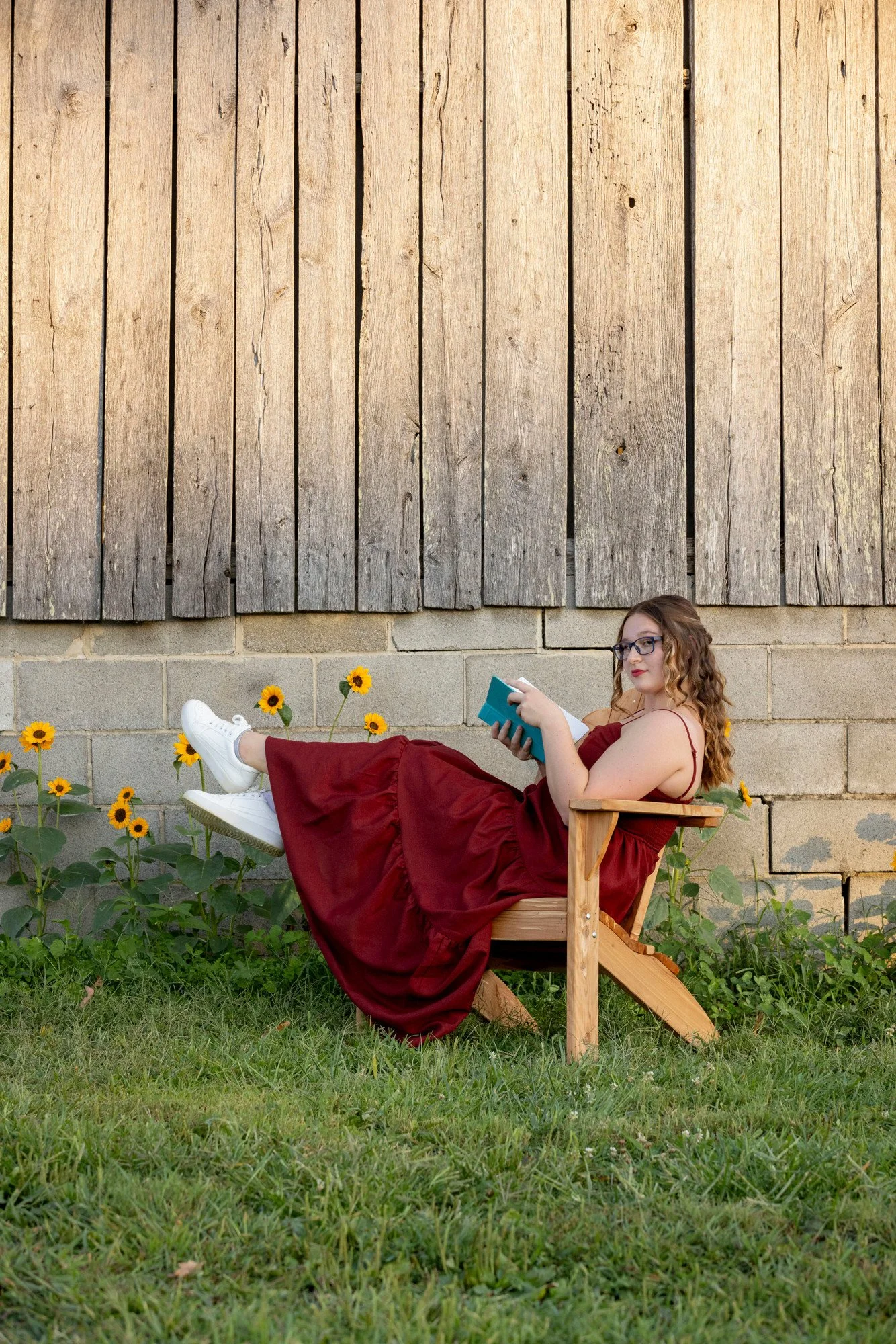 A woman with red hair, glasses, and a red dress sitting in a wooden chair in a garden with yellow flowers, reading a book, with a wooden wall in the background.