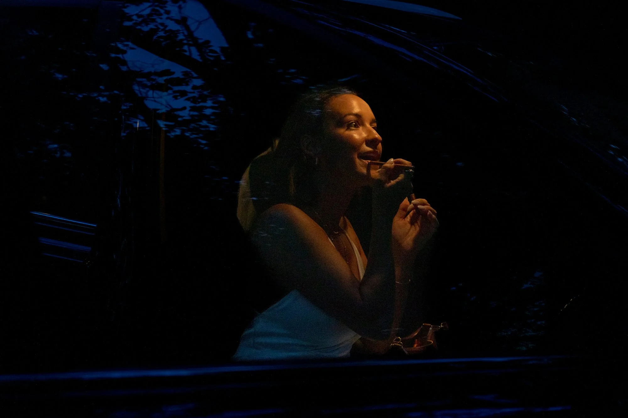 A woman with brown hair smiling and holding a drink, seen through a dark window with reflections of trees and the night sky.