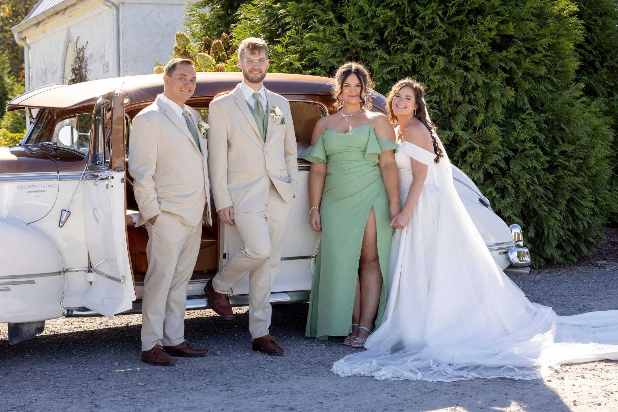 A group of five people dressed in wedding attire standing next to a vintage white car with a brown roof, outdoors in front of green bushes and a white building, smiling and posing for a photo.