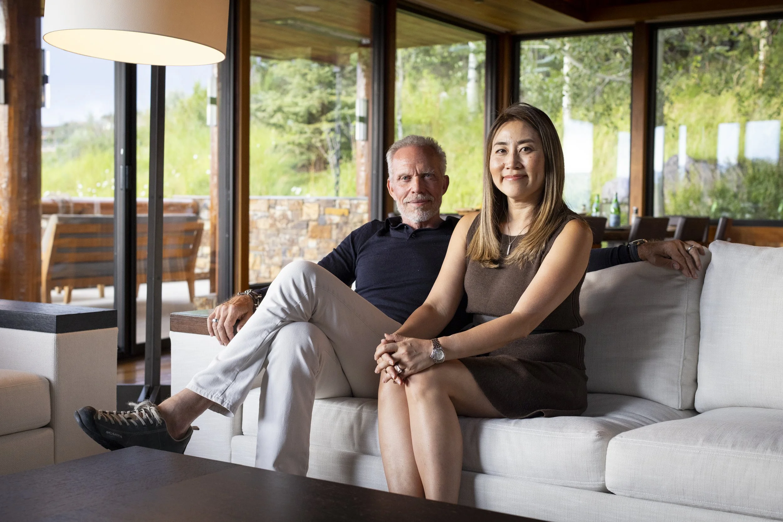 A man and a woman sitting on a white sofa in a room with large windows showing greenery outside, with the man wearing a black shirt and light-colored pants, and the woman wearing a brown sleeveless top and dark skirt.