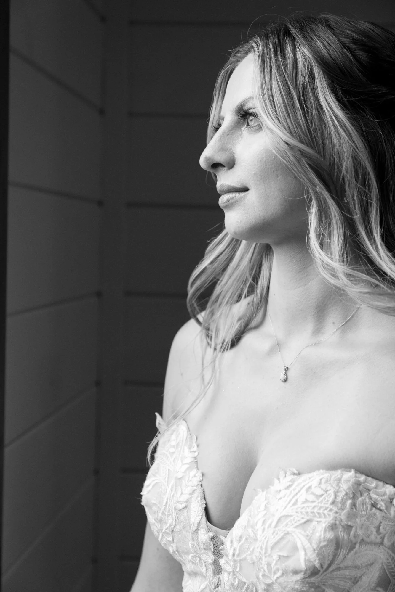 Black and white photo of a woman with light hair, wearing a lace dress and a small pendant necklace, looking to the side.