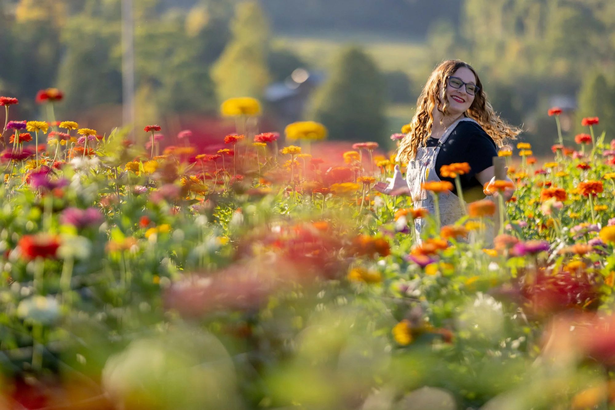 A young woman with curly red hair and glasses smiling while standing in a colorful flower field with yellow, red, and purple flowers, sunny weather, and trees in the background.