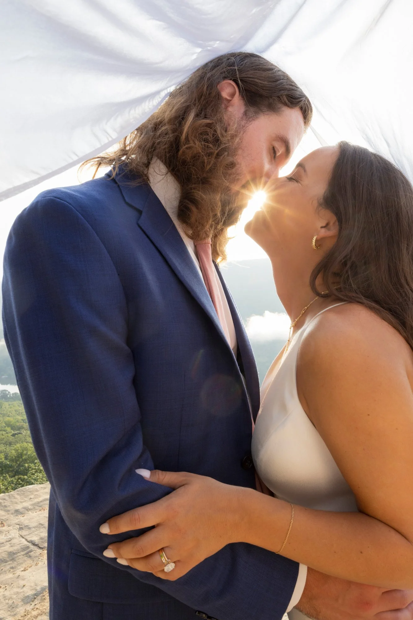 A man and woman sharing a kiss, standing outdoors with sunlight shining between them, with trees and sky in the background.