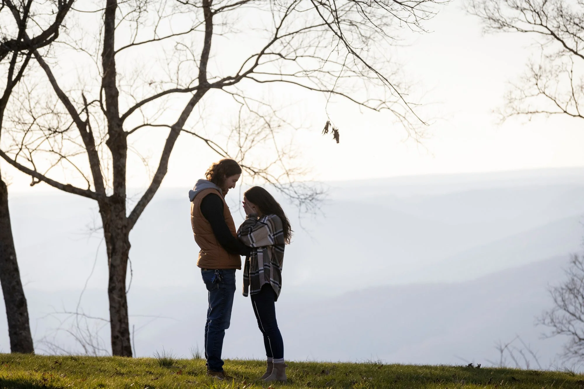 A couple standing close together outdoors near a leafless tree, with their foreheads touching and hands clasped, under an overcast sky, during winter.