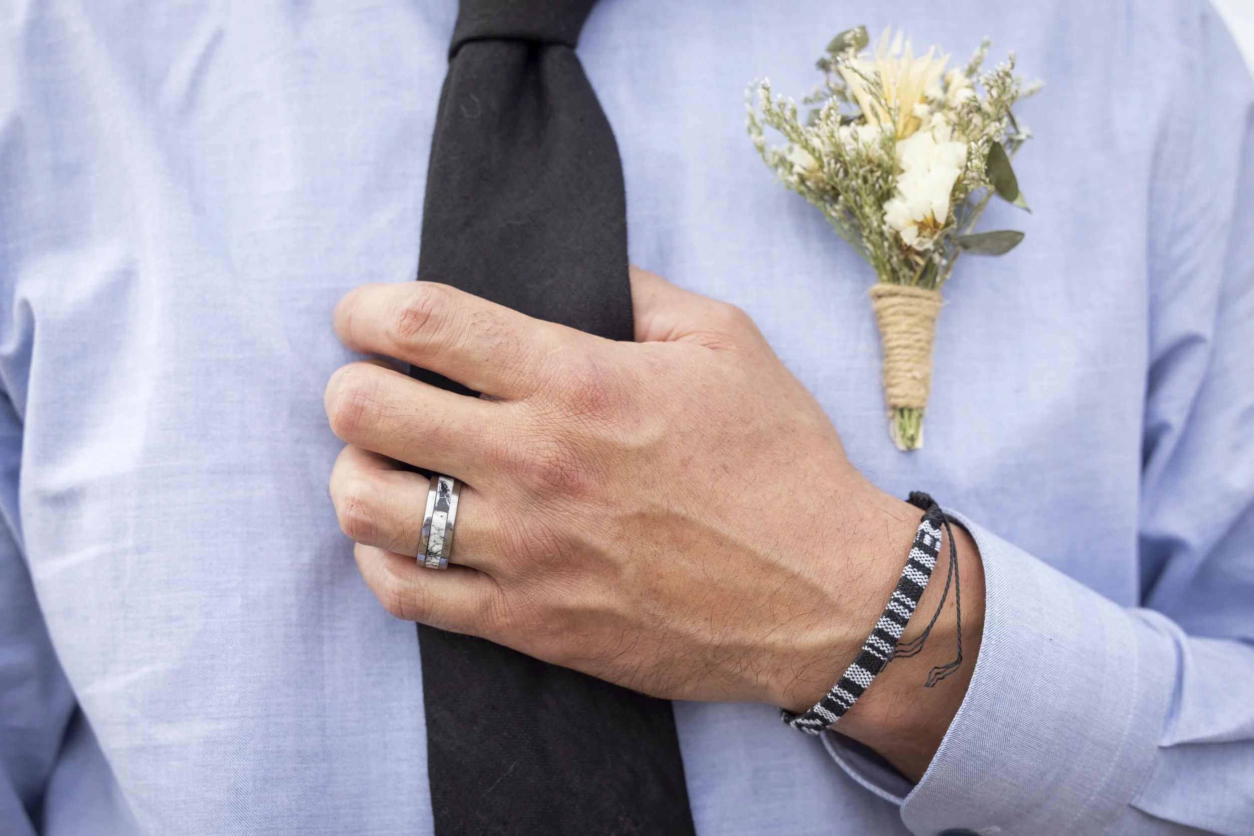 Person wearing a light blue shirt with a boutonniere of white flowers and greenery attached to the lapel, holding a black object close to their chest, with a silver ring on their finger and several black and white bracelets on their wrist.
