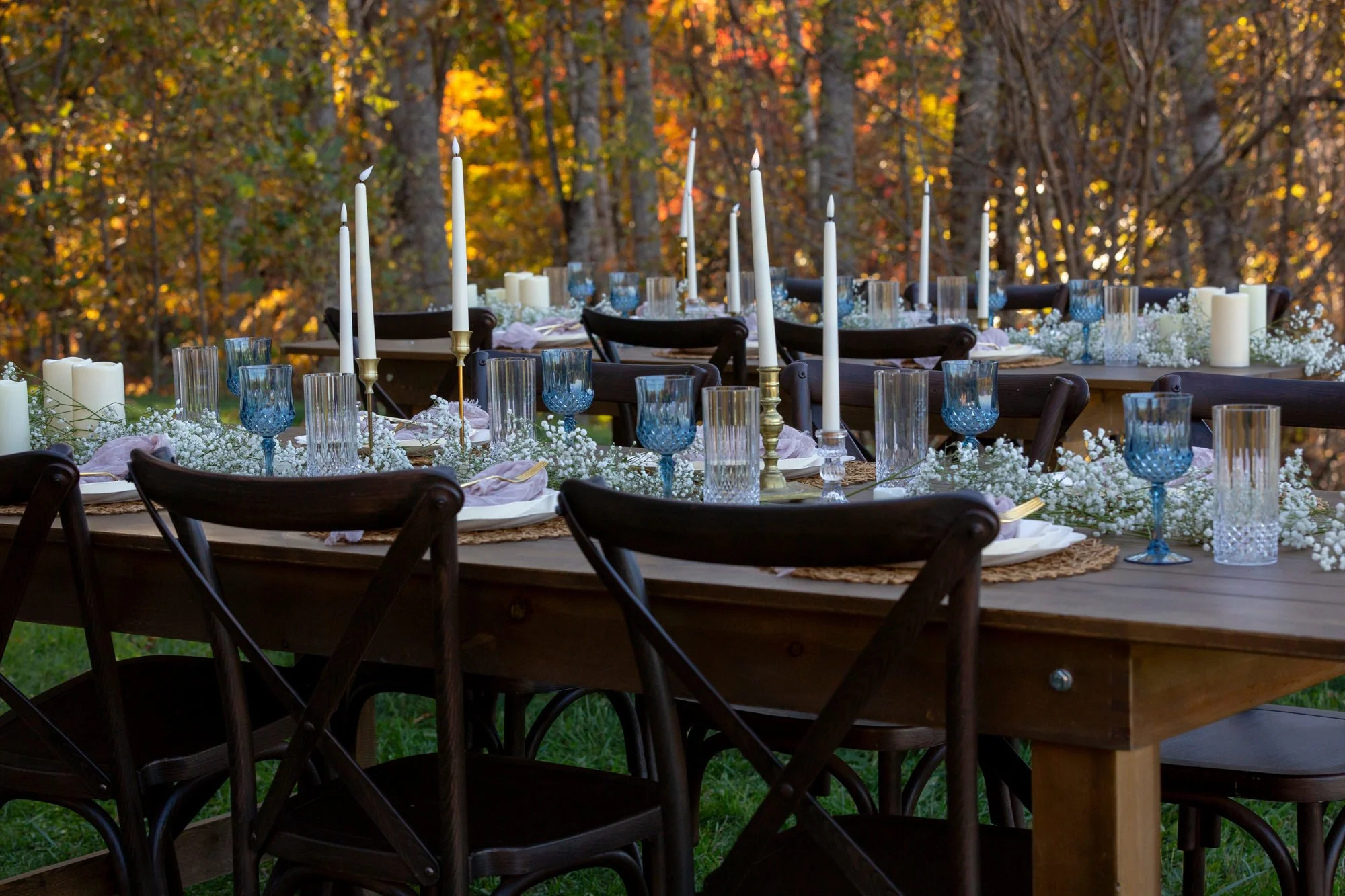 Outdoor dining table decorated with white flowers, candles, and blue glasses, set against a forest backdrop during fall with colorful trees.