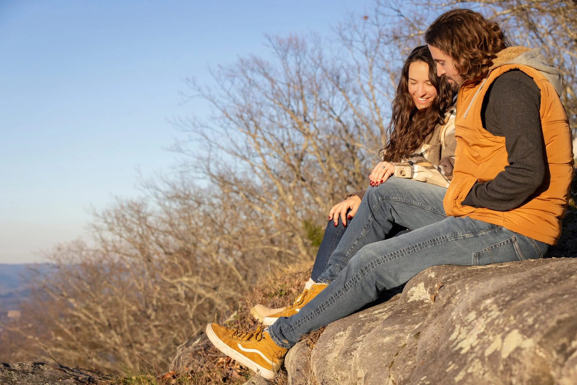 A man and a woman sitting on a large rock outdoors, smiling and looking at each other. The woman has long curly brown hair and is wearing a beige jacket, while the man has long wavy brown hair and a beard, wearing a mustard yellow vest over a black s