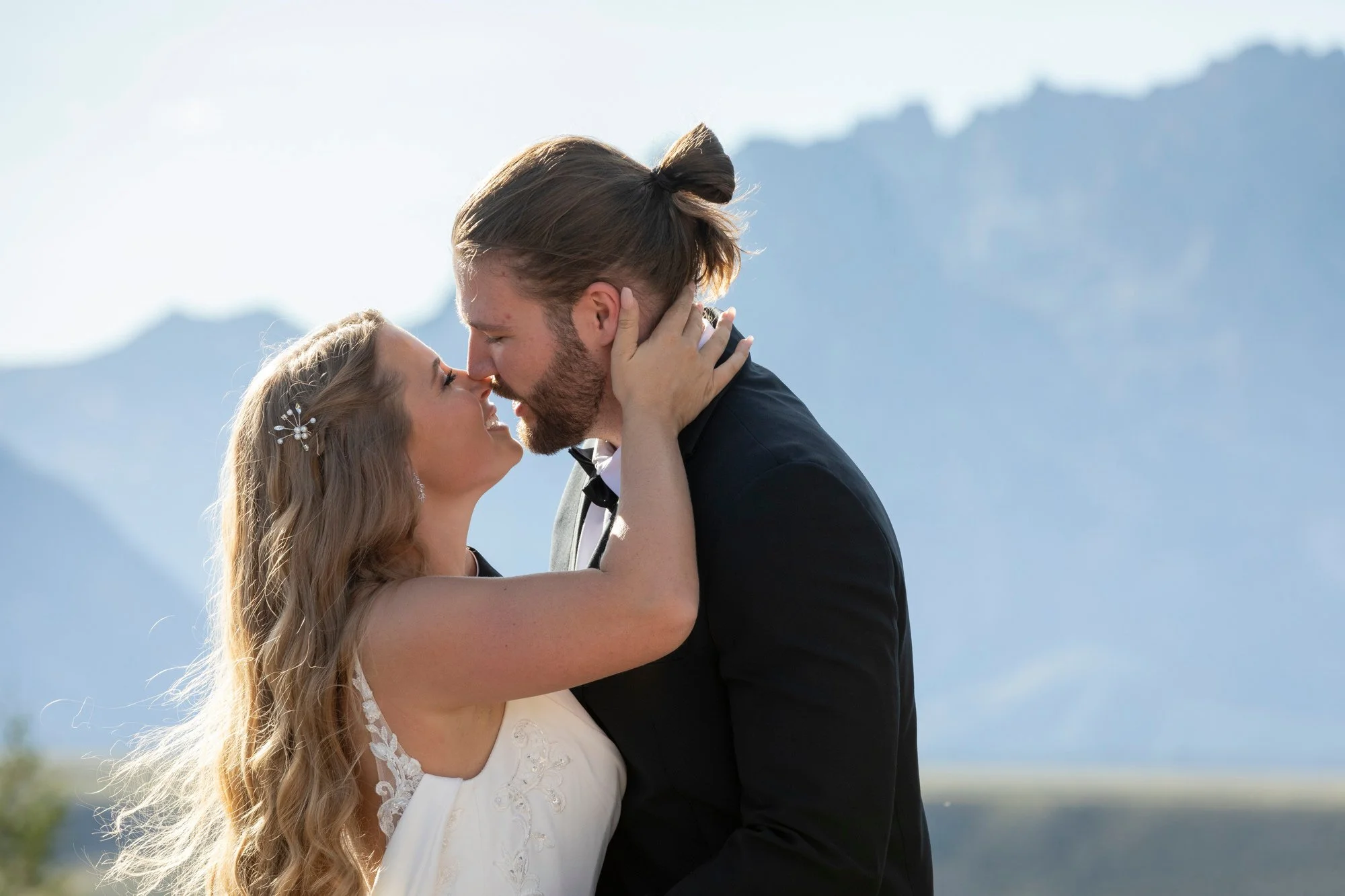 A couple dressed in wedding attire sharing an intimate moment outdoors with a mountain range in the background.