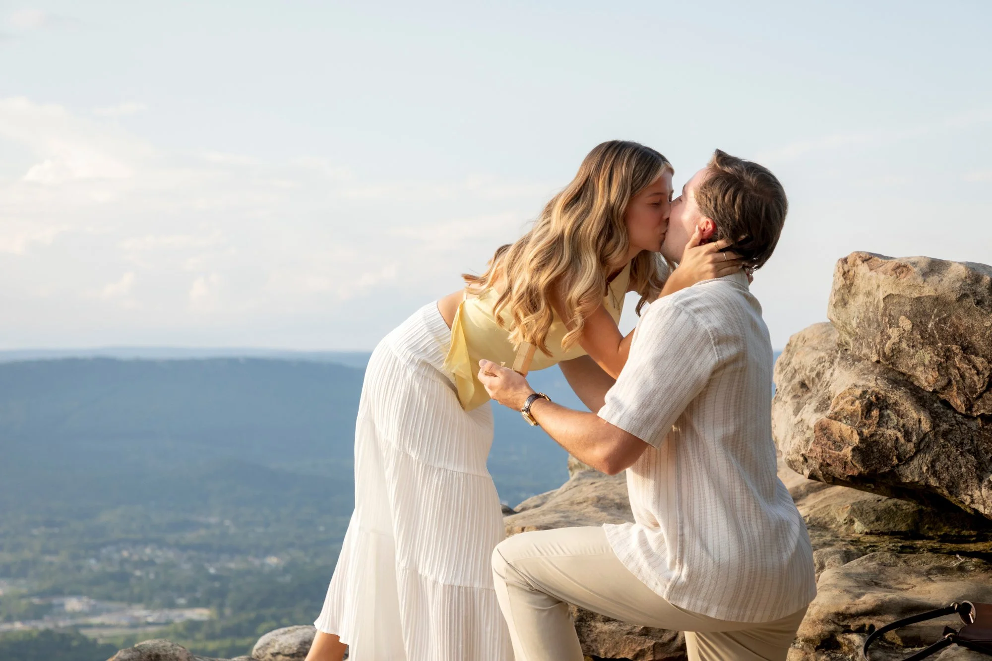 A couple shares a kiss on a mountain with a view of a valley and sky in the background.