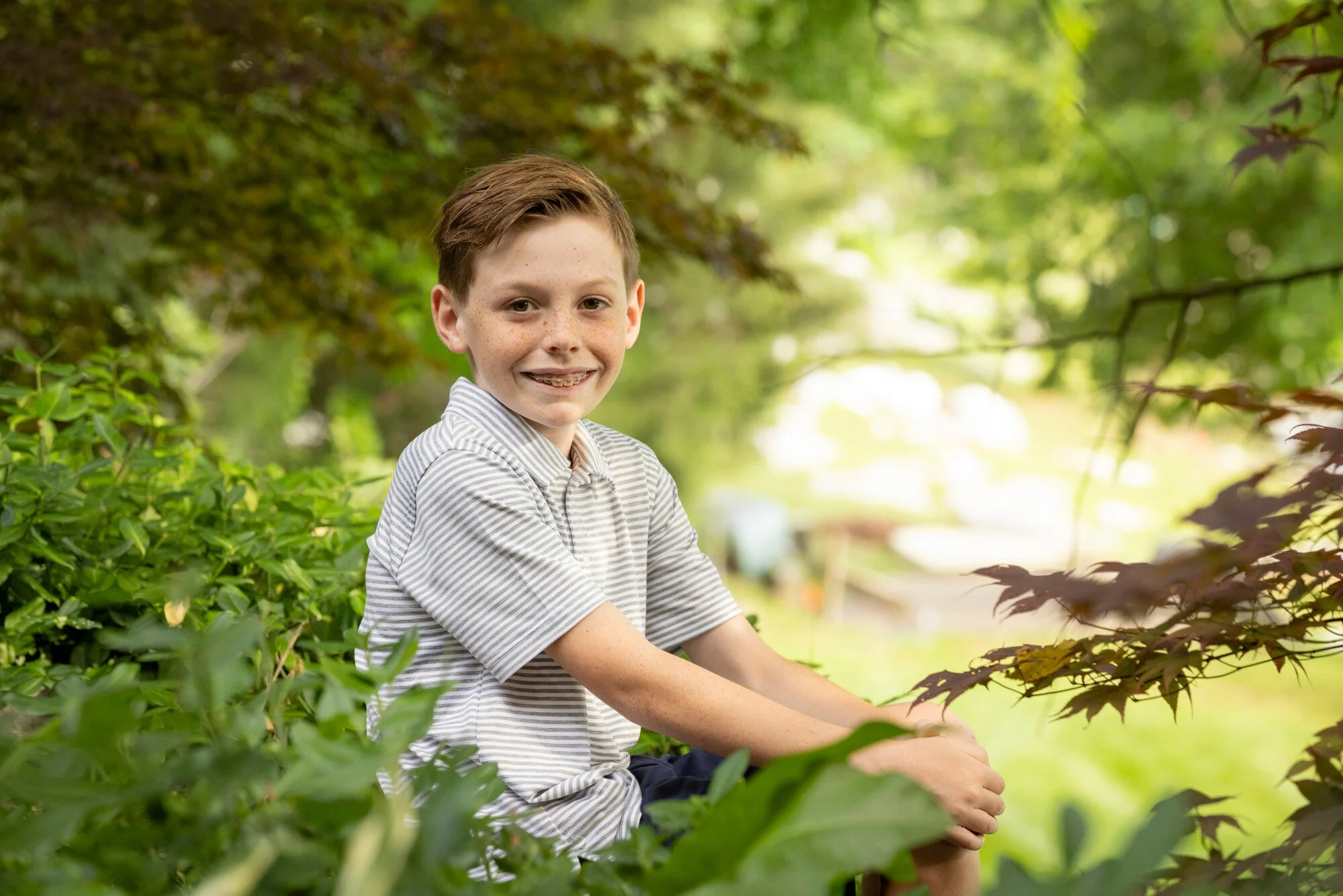 A young boy with short brown hair, braces, and freckles smiling wearing a striped shirt, sitting outdoors among green bushes and trees on a sunny day.
