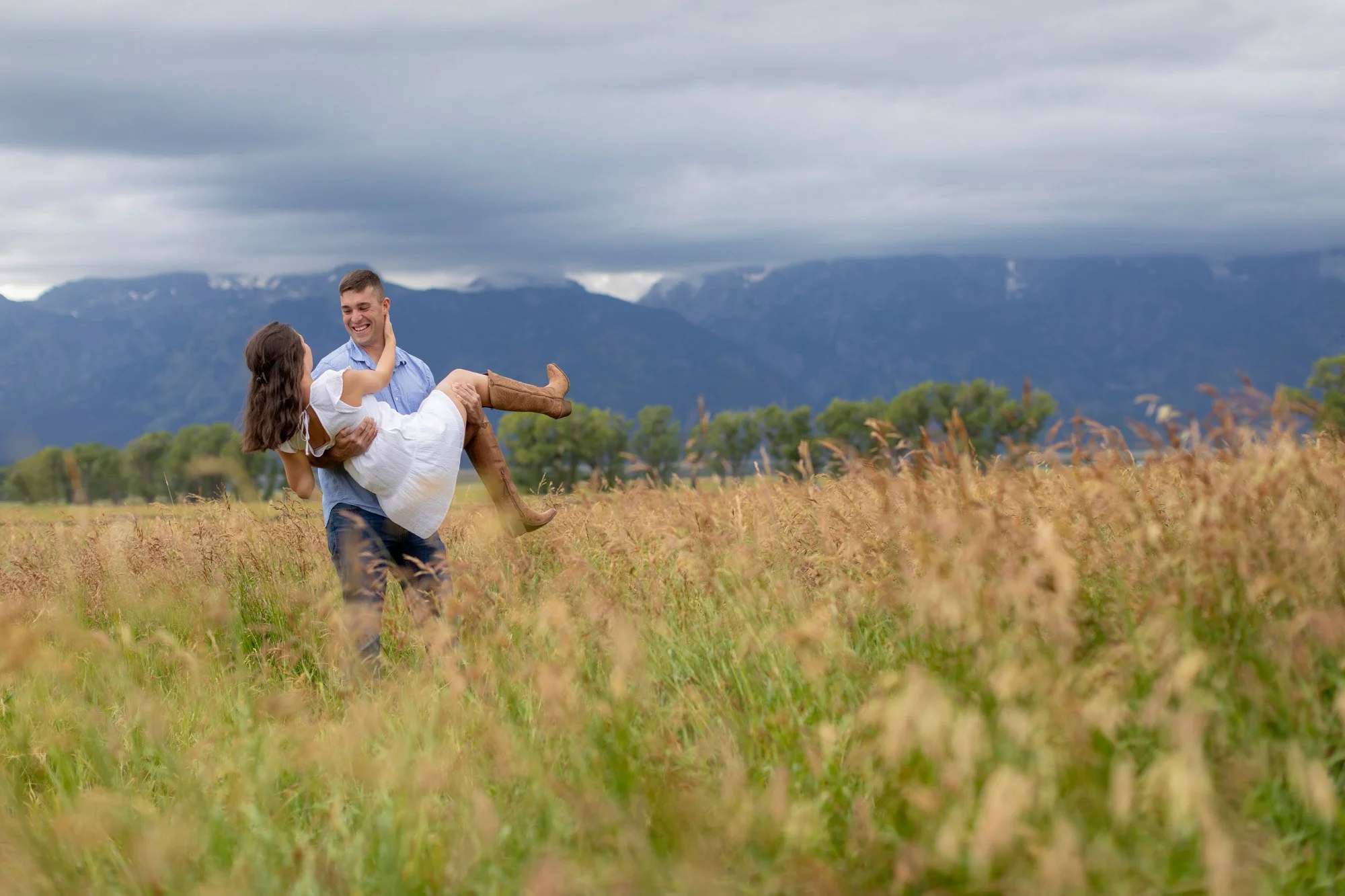 A man is carrying a woman in a white dress and brown boots in a field of tall grass, with mountains and a cloudy sky in the background.