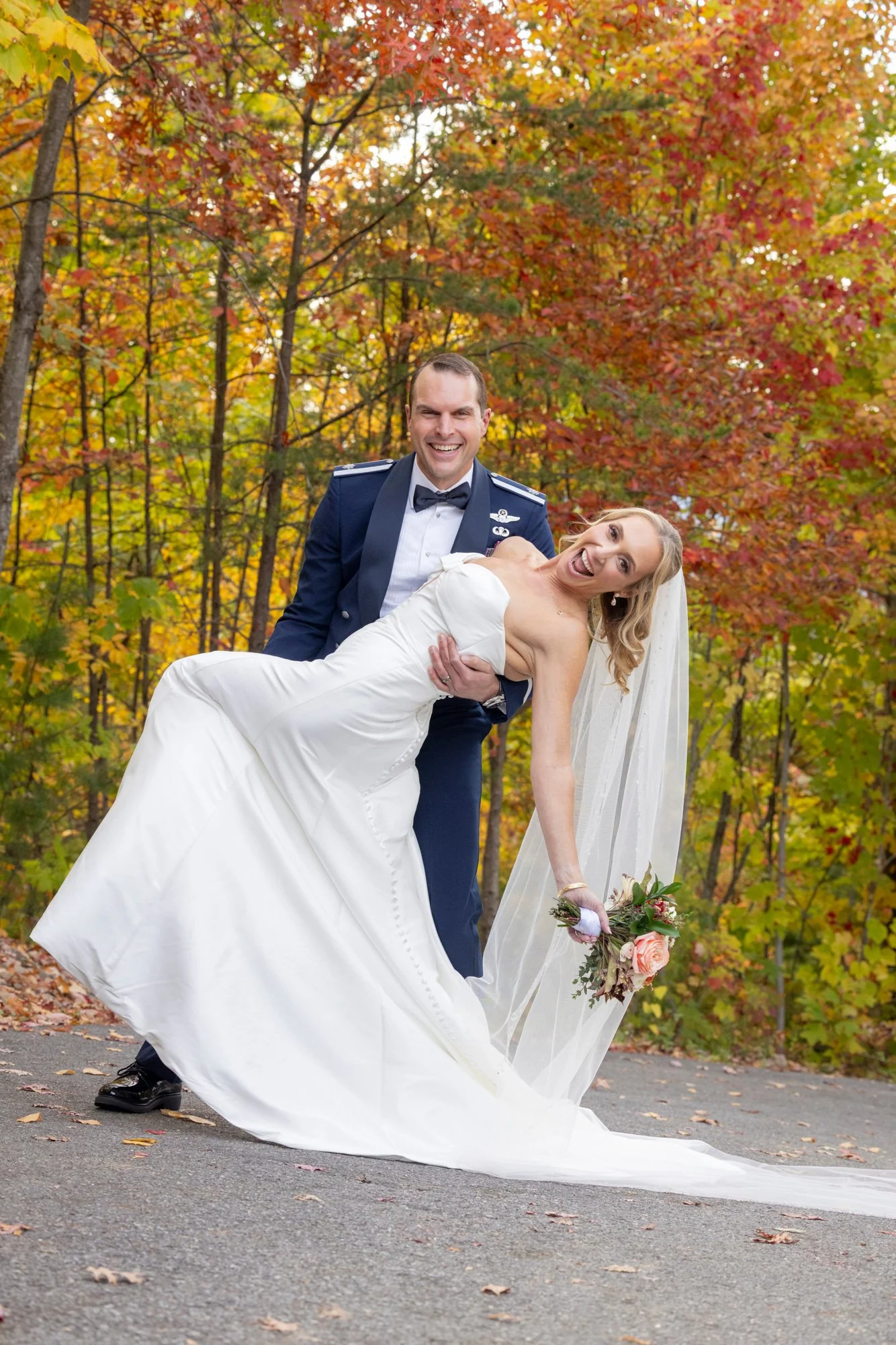 A smiling groom in a navy blue military uniform holding a laughing bride in a white wedding dress outdoors with autumn leaves in the background.