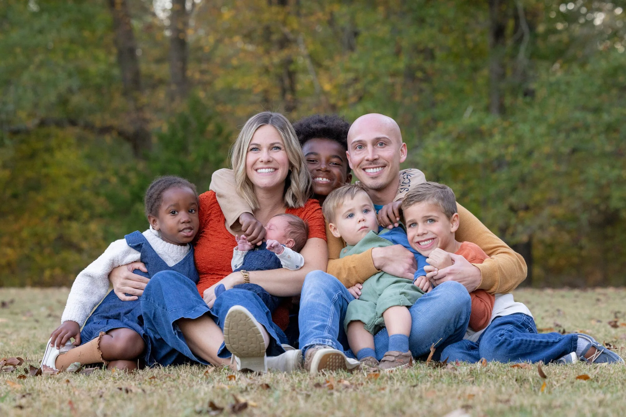 A happy multiracial family of seven, including adults and children, sitting together outdoors in a park surrounded by fall foliage, smiling and embracing each other.
