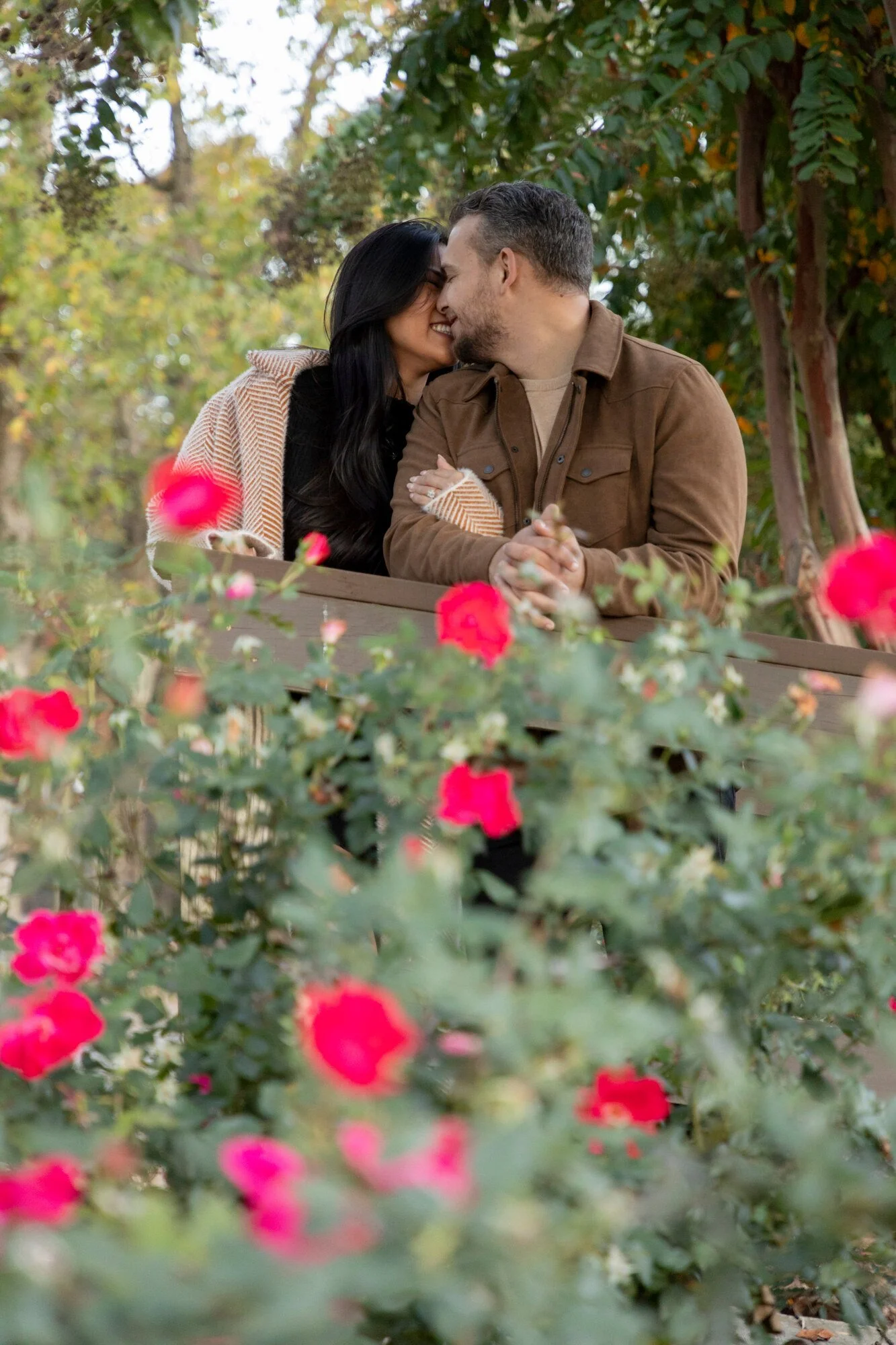 A couple sitting by a wooden fence outdoors, surrounded by pink flowers and green trees, sharing a close moment with their foreheads touching.