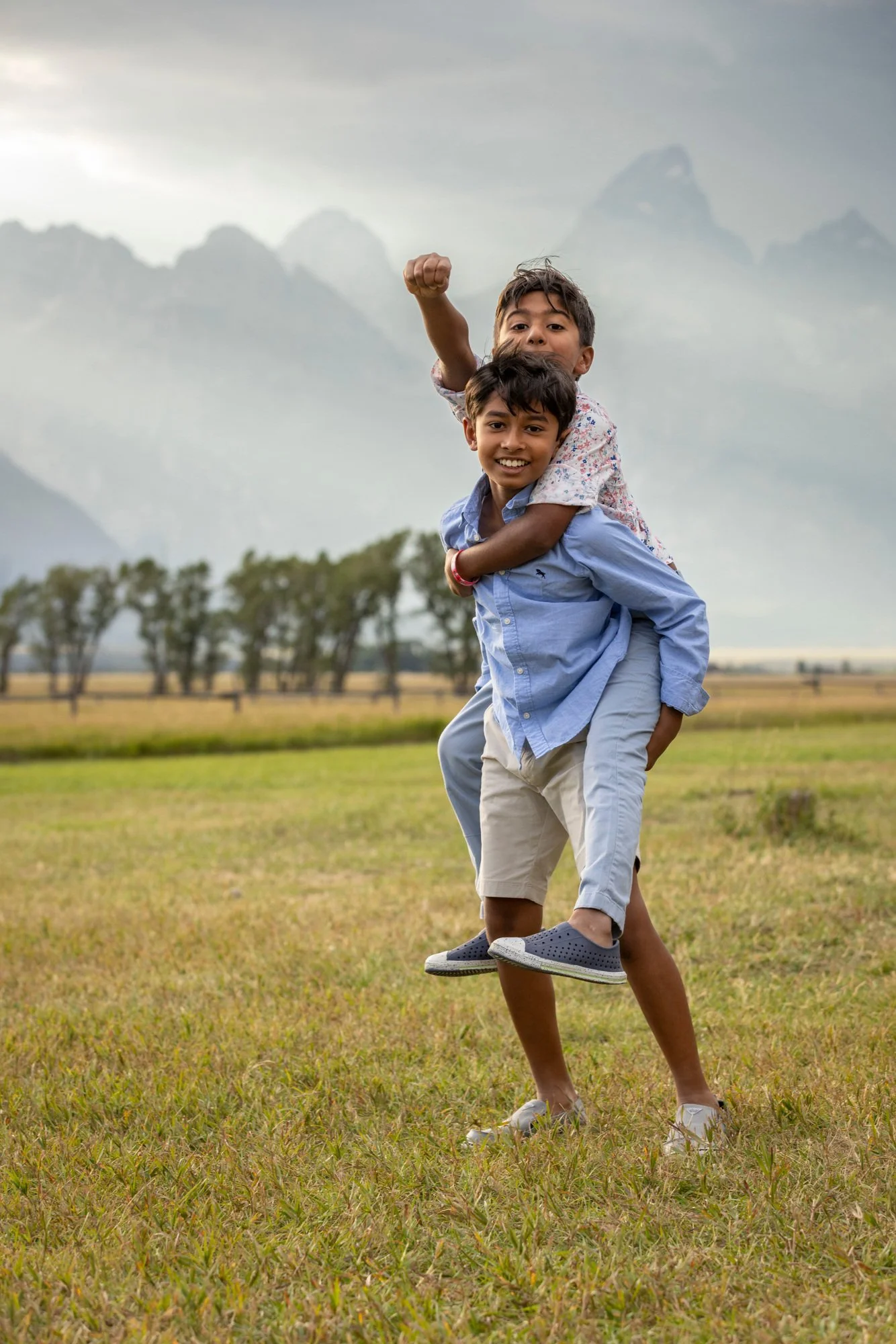 Two young boys playing in a grassy field with mountains and cloudy sky in the background. One boy gives the other a piggyback ride, and both are smiling.