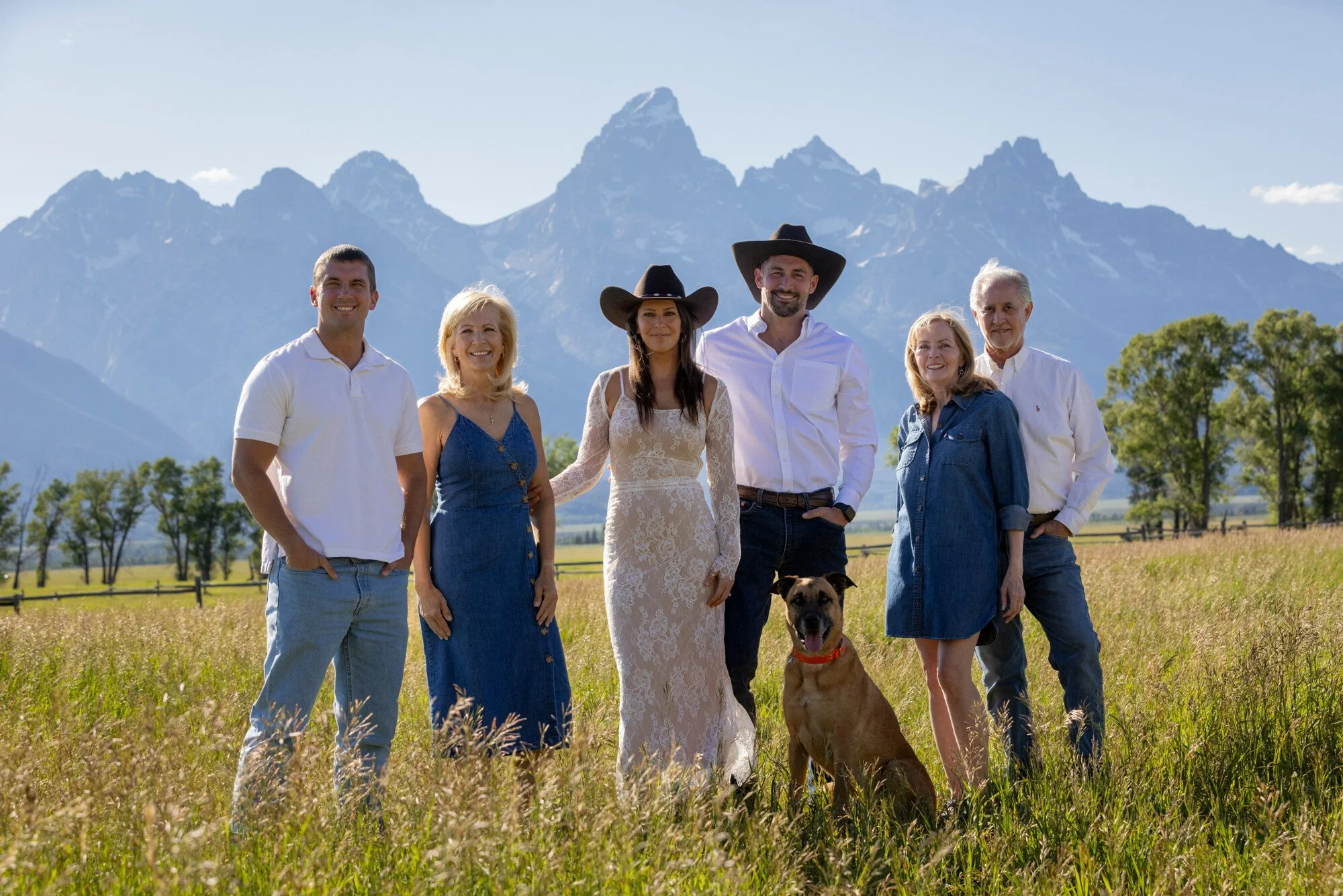 Group of six people standing in a field with mountains in the background, including a woman in a lace dress and two people wearing cowboy hats, along with a brown dog.