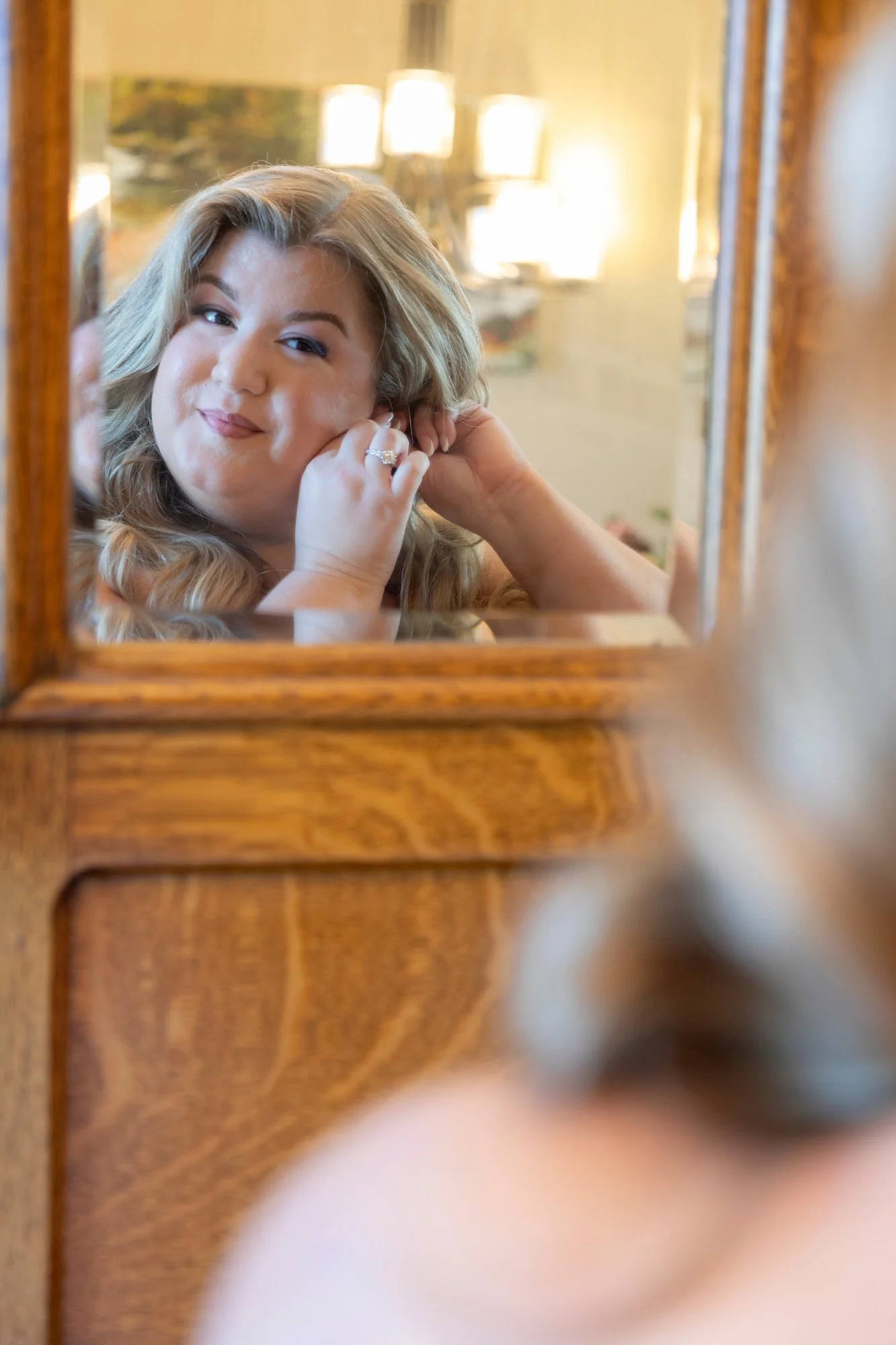 A woman with blonde hair looking into a mirror while adjusting her earring in a warmly lit room.