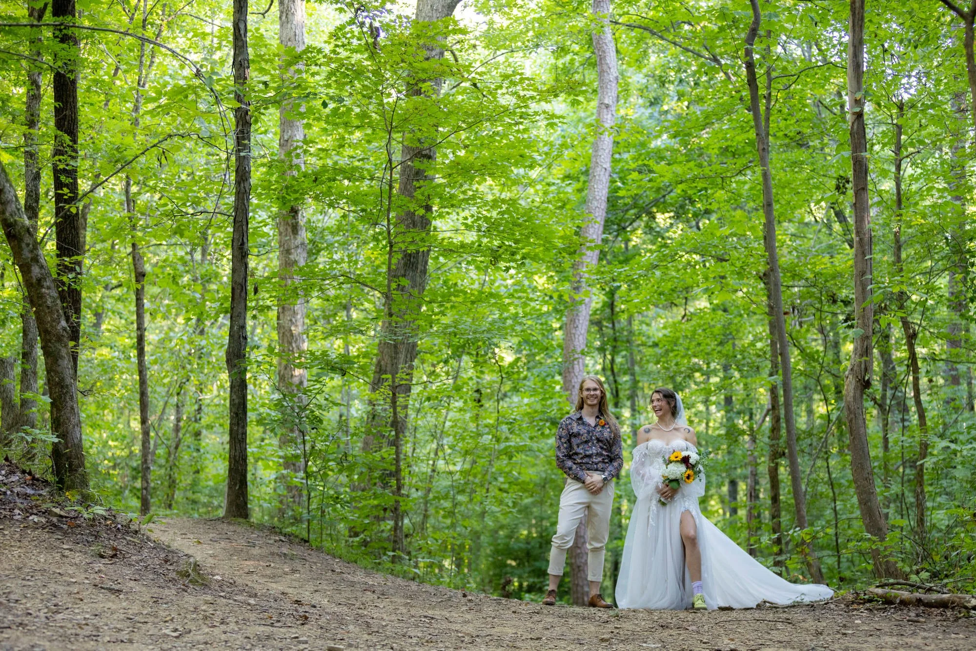 A couple dressed in wedding attire walking through a lush green forest with tall trees and dense foliage. The bride is holding a bouquet of sunflowers and wearing a white wedding dress with a train, while the groom is wearing a patterned shirt and li