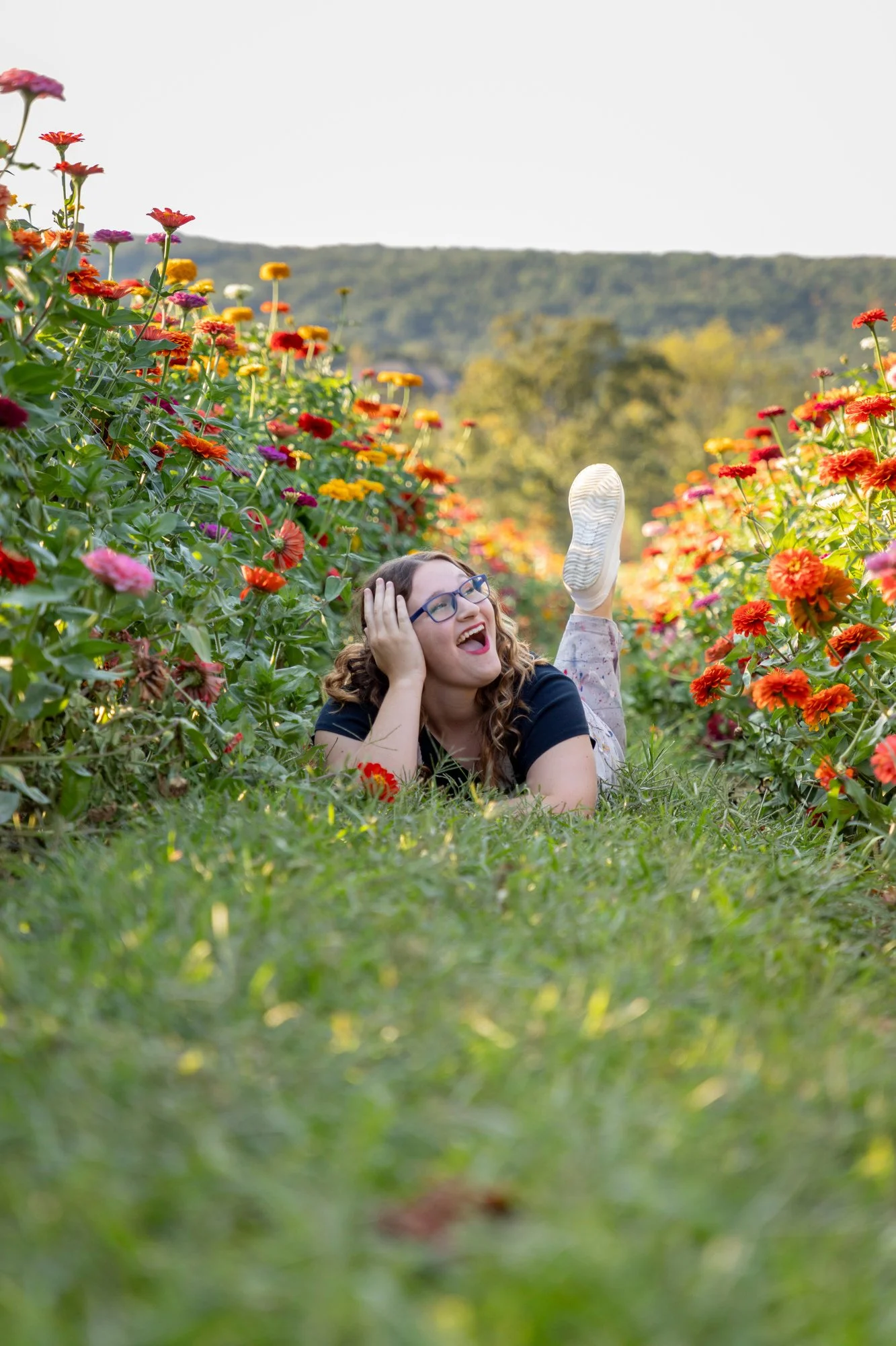 A woman lying on the grass in a colorful flower garden, smiling and wearing glasses, with her head resting on one hand and her leg raised in the air.