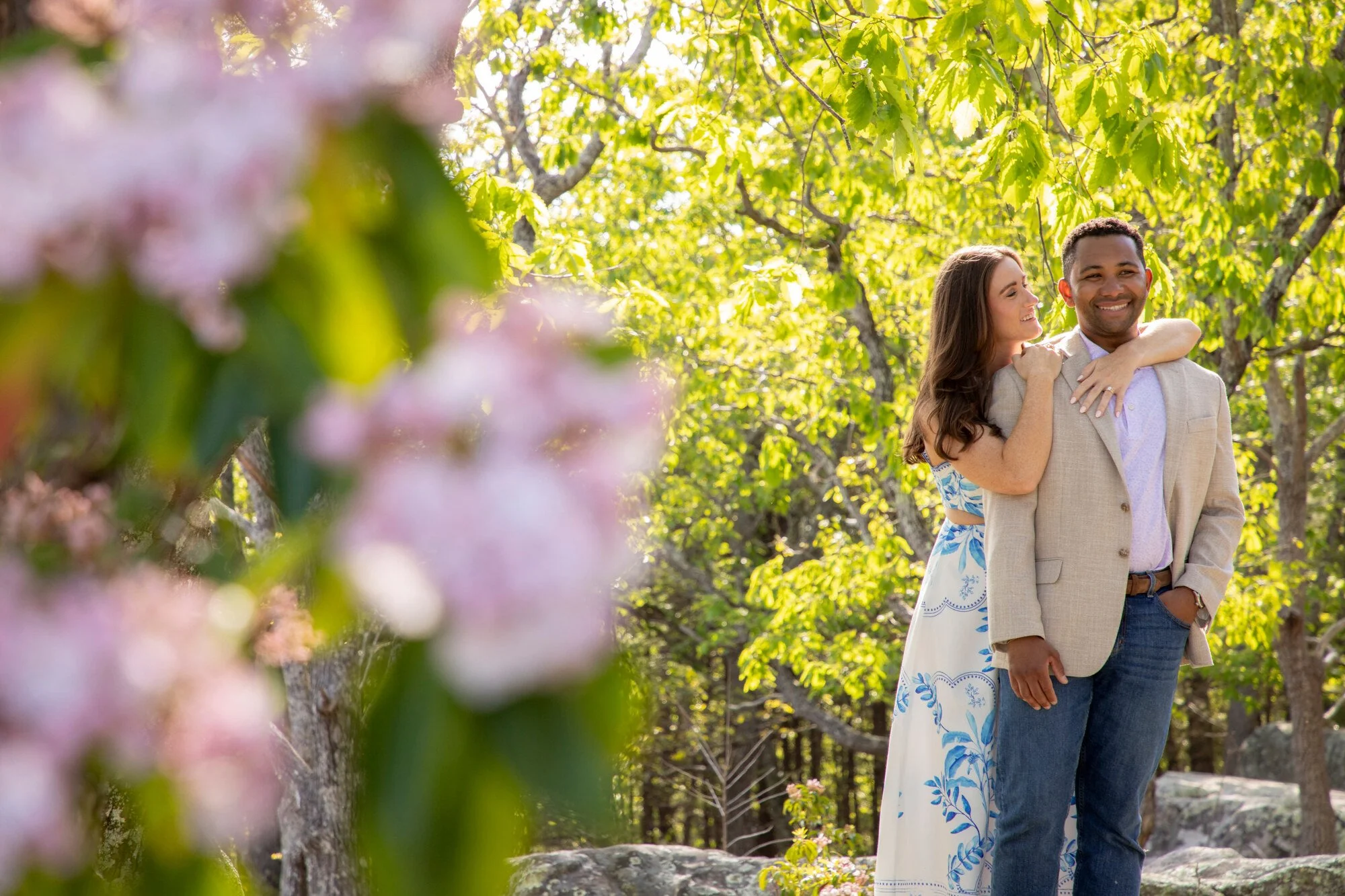 A smiling woman and man in a park surrounded by green trees, with pink flowers in the foreground.