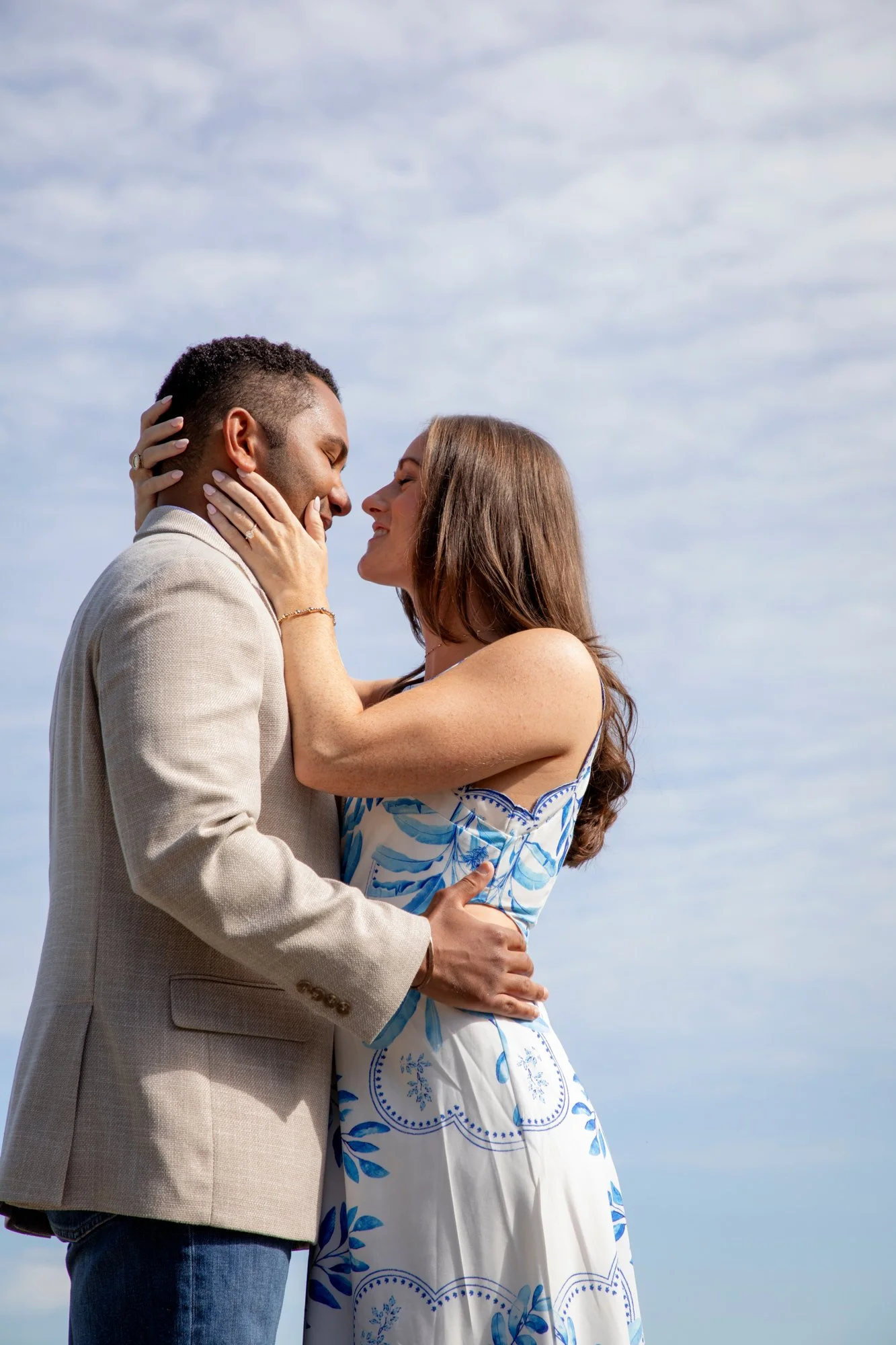 A couple standing close together outdoors, smiling and touching faces, with cloudy sky in the background.