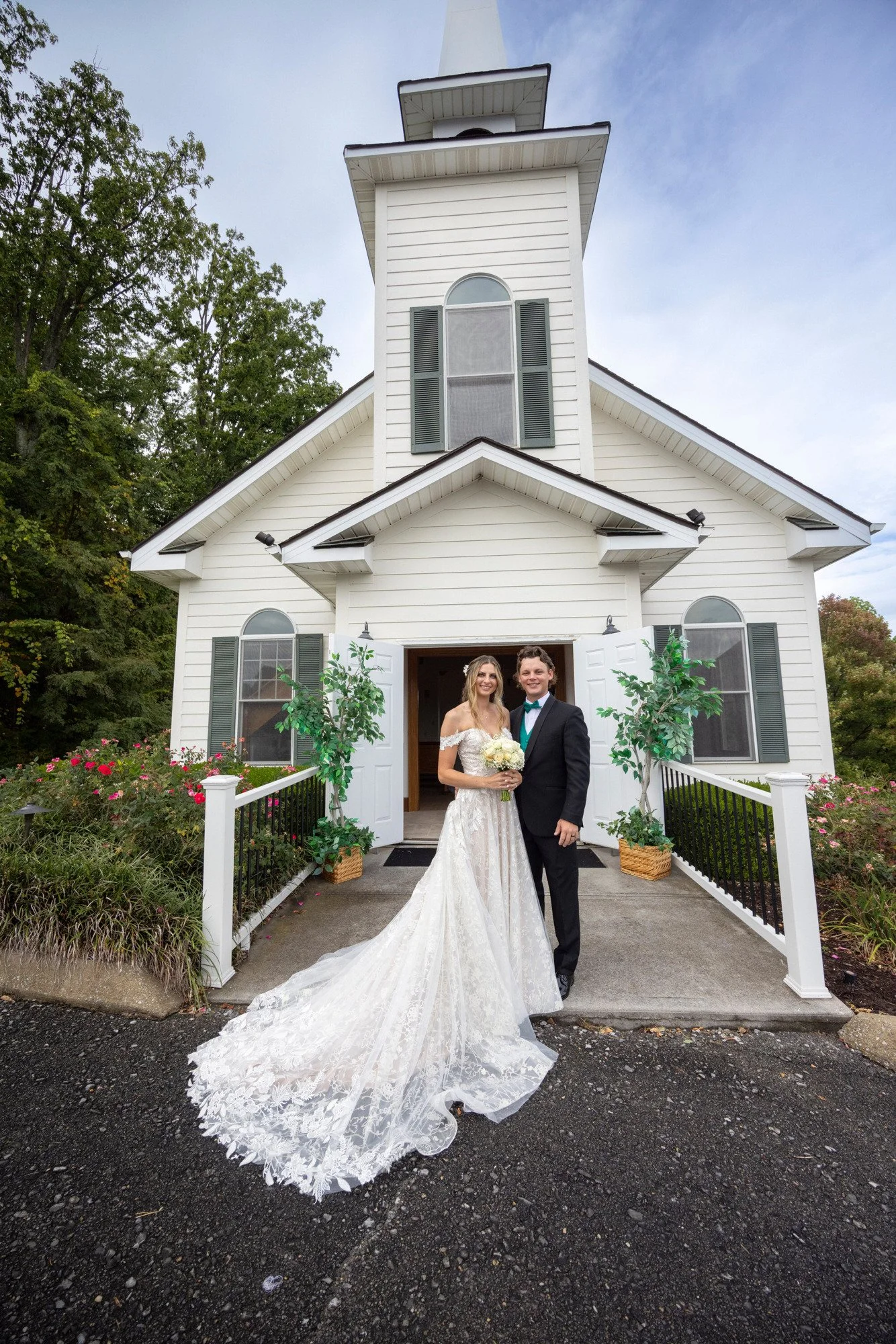 A bride and groom standing in front of a white church with open double doors, surrounded by greenery and flowers, on a cloudy day.