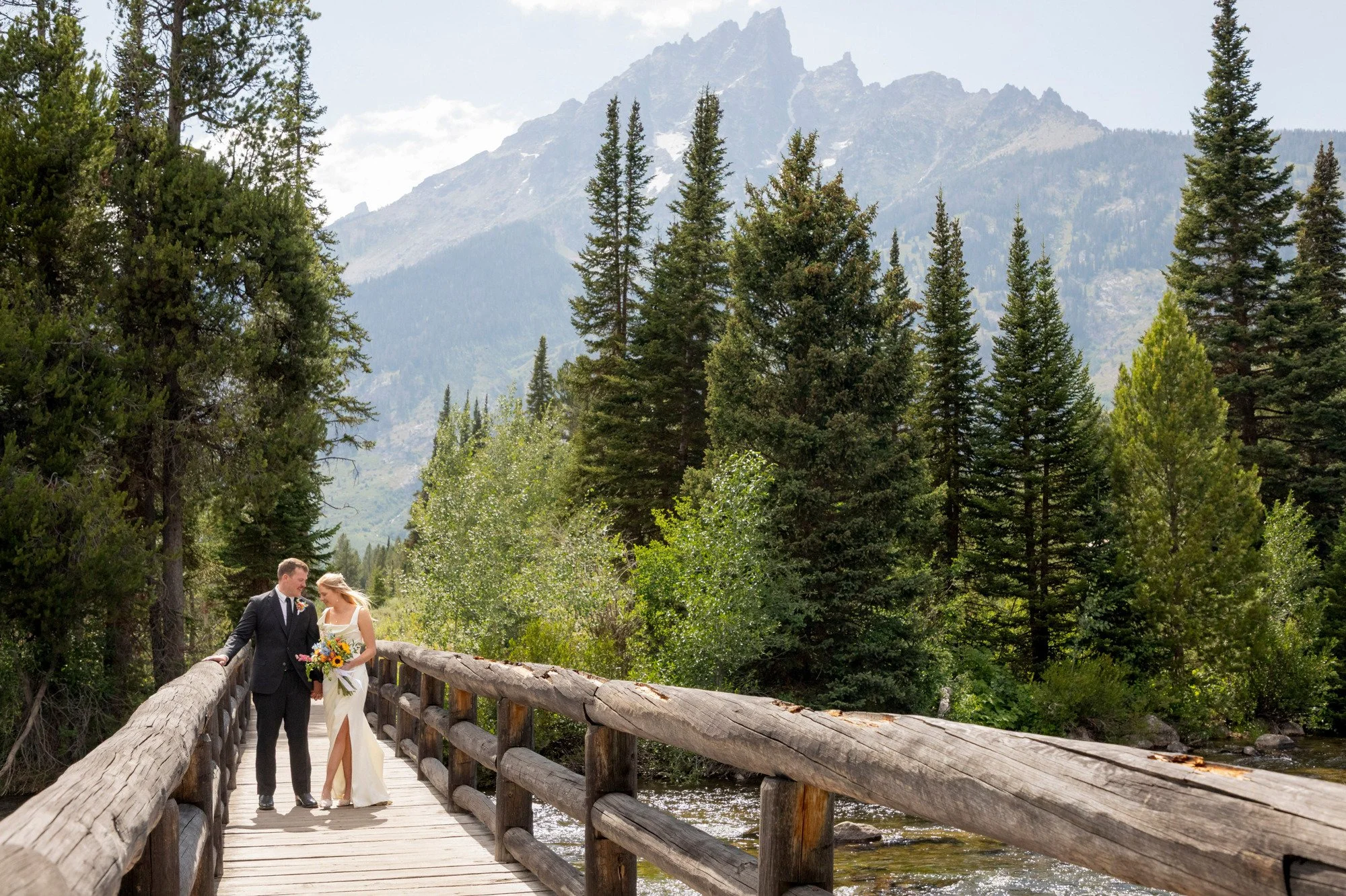 A bride and groom walking on a wooden bridge over a stream in a forested mountain area, with tall pine trees and mountain peaks in the background.